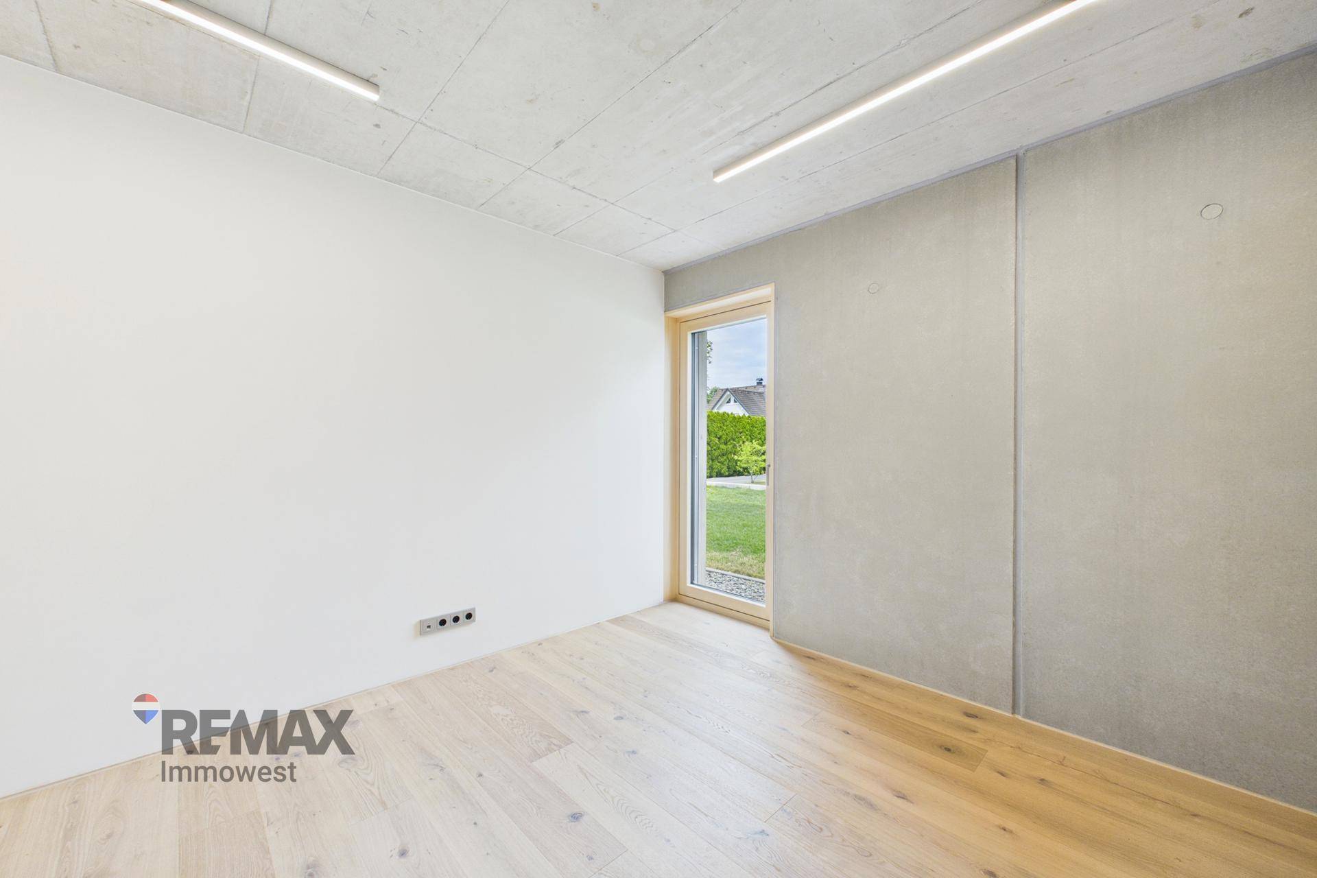 Modernes Zimmer mit Holzboden, Sichtbetondecke und Fenster zum Garten.