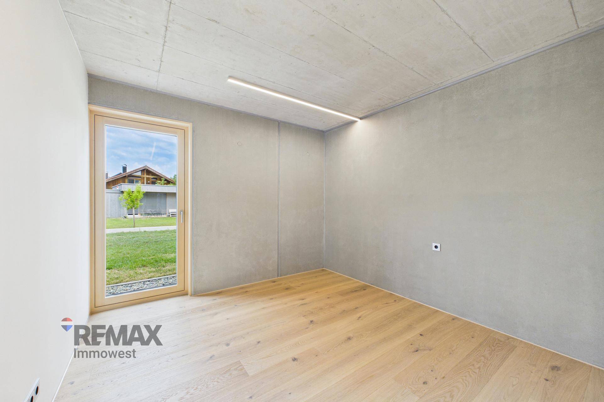 Geräumiges Zimmer mit Holzboden, Sichtbetondecke und Blick ins Grüne.