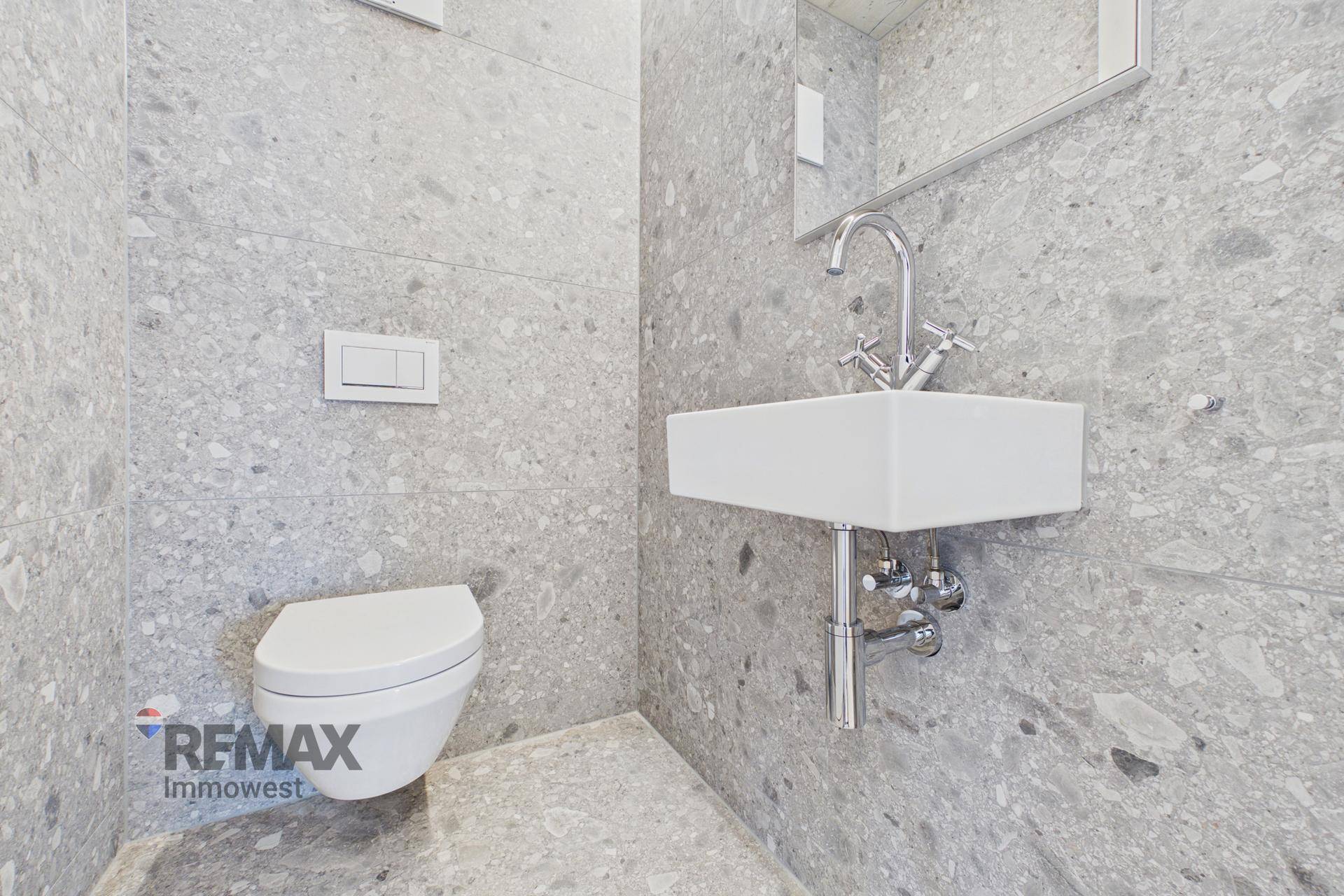 Modernes Gäste-WC mit grauen Fliesen, wandhängendem WC und kleinem Waschbecken.