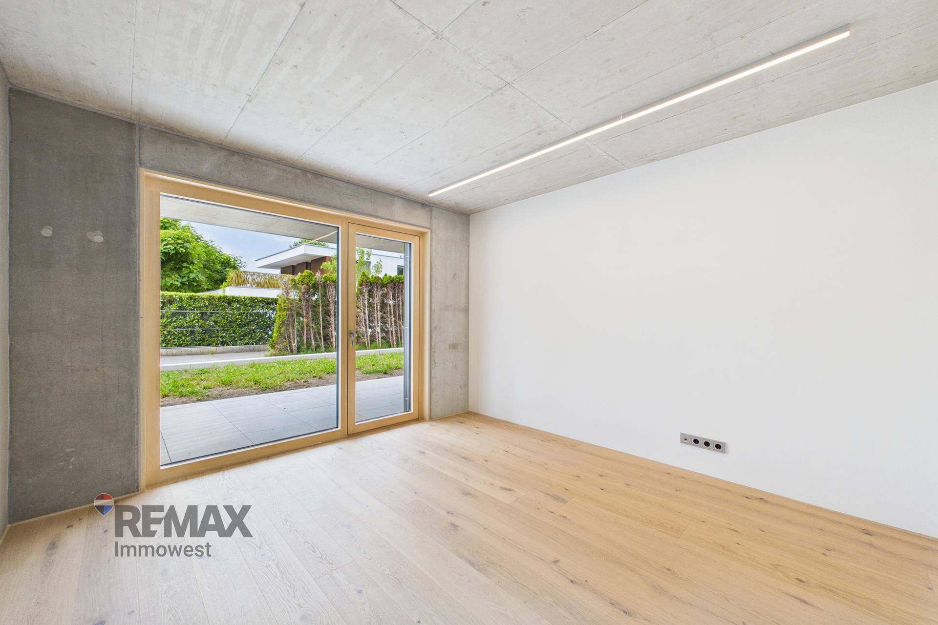 Geräumiges Zimmer mit Holzboden, Sichtbetondecke und bodentiefen Fenstern zum Garten.