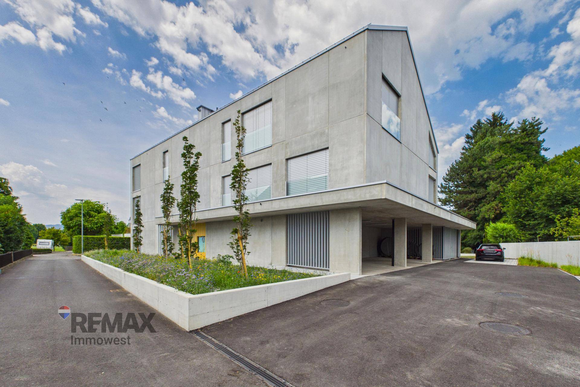 Modernes Mehrfamilienhaus mit Betonfassade, großen Fenstern und Zufahrt zu den Parkplätzen.