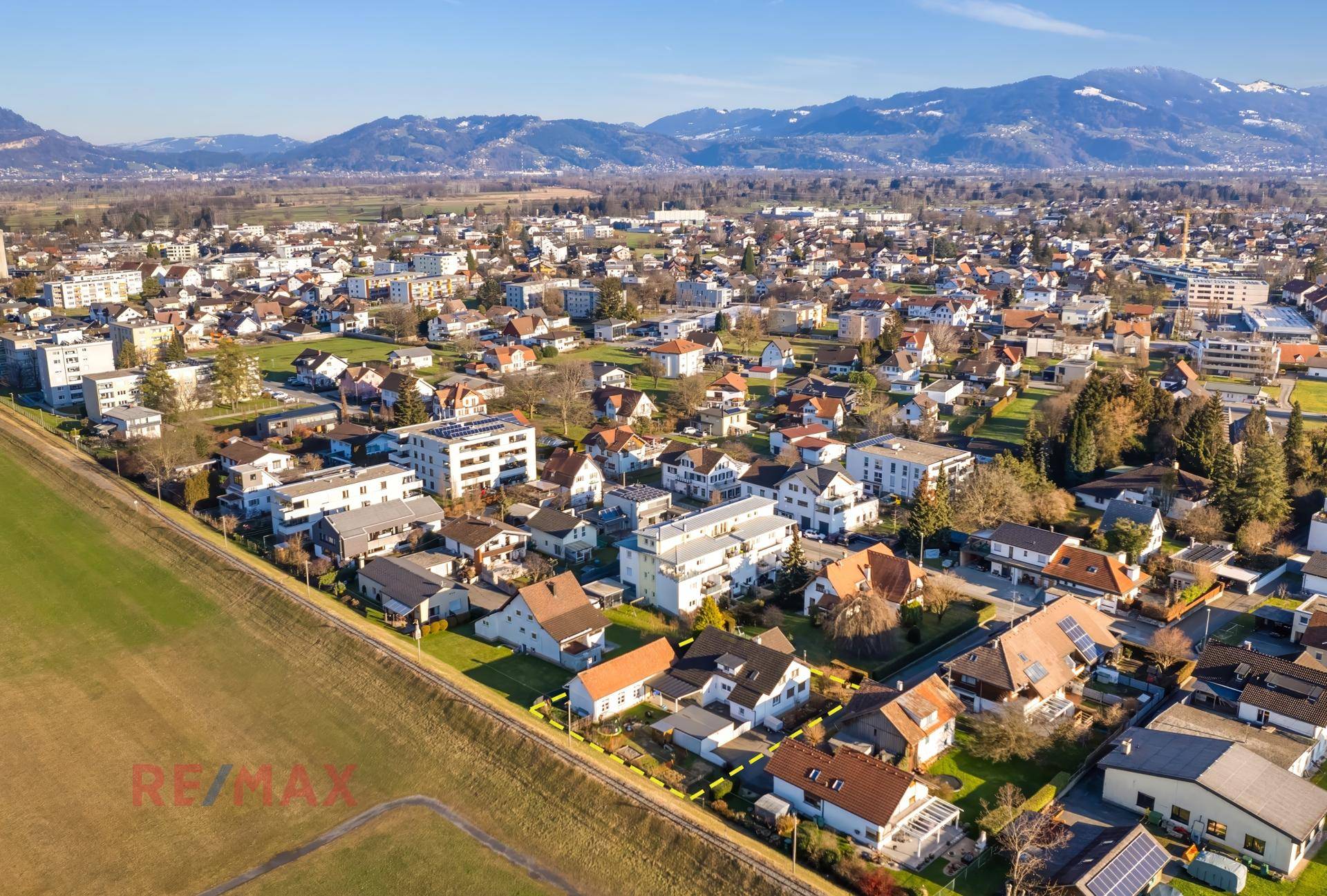 Weitläufige Luftaufnahme einer Wohnsiedlung mit grünen Feldern und Bergen am Horizont.