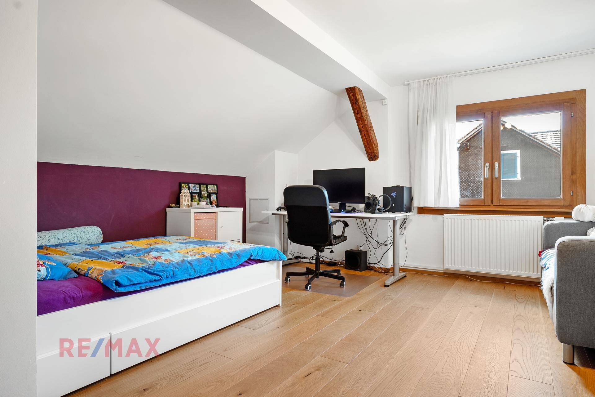 Kinderzimmer mit Holzboden, lila Akzentwand, Bett und Schreibtisch am Fenster.