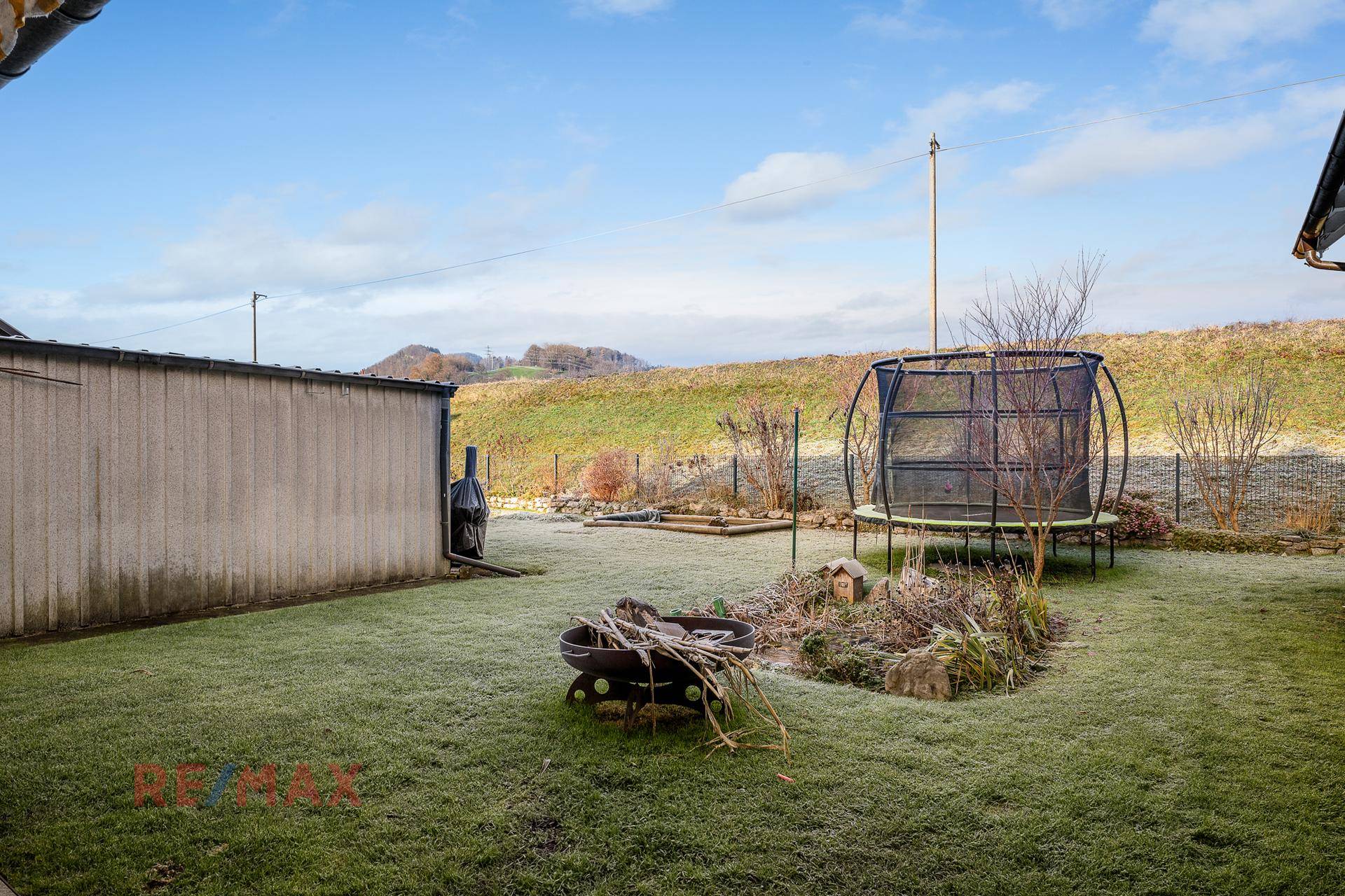 Großer Garten mit Rasenfläche, Trampolin und Feuerstelle, umgeben von Hügeln.