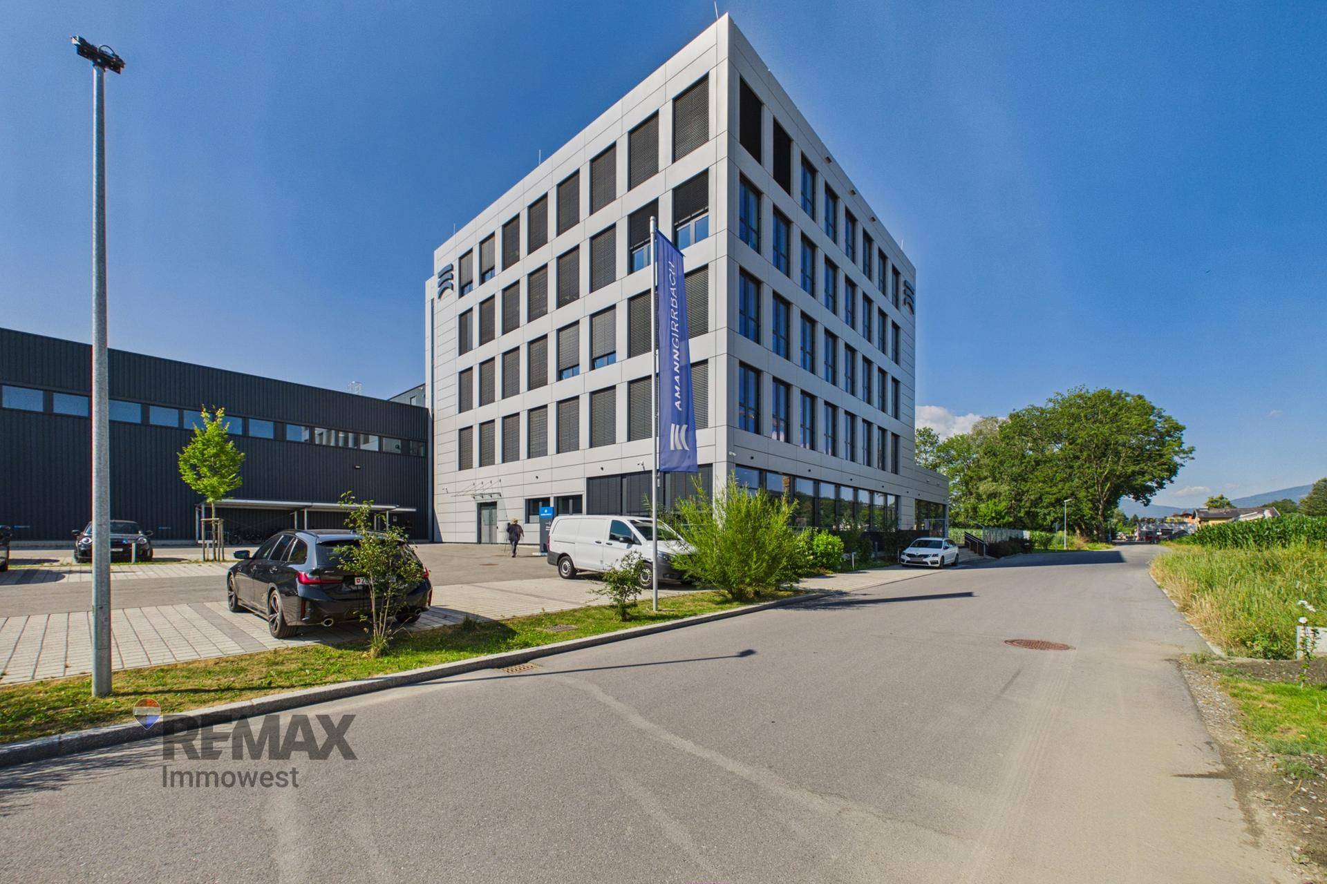 Modernes Bürogebäude mit heller Fassade und vielen Fenstern, umgeben von Parkplätzen und Grünflächen.