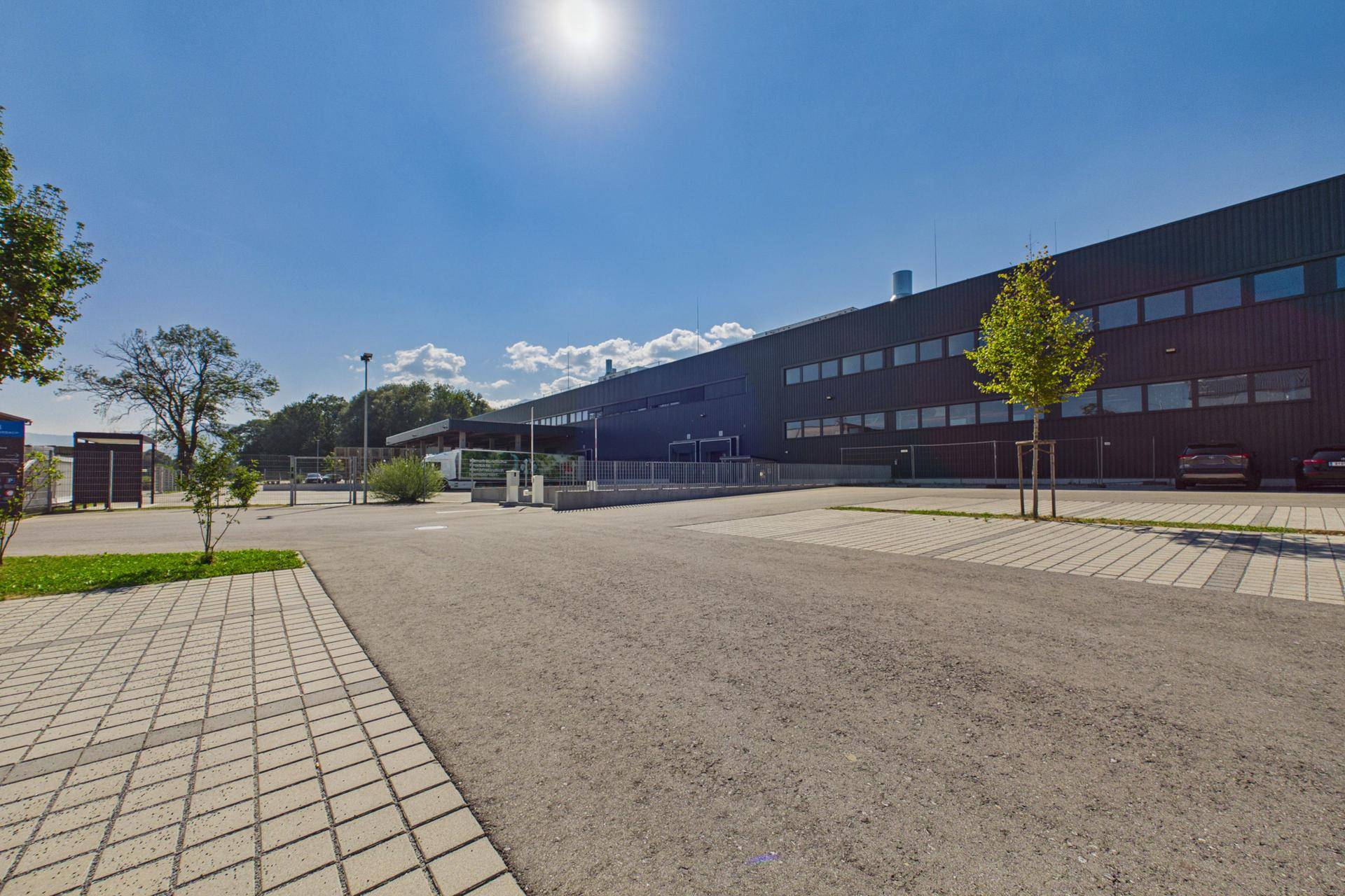 Modernes Industriegebäude mit dunkler Fassade, vielen Fenstern und großem gepflastertem Parkplatz unter blauem Himmel.