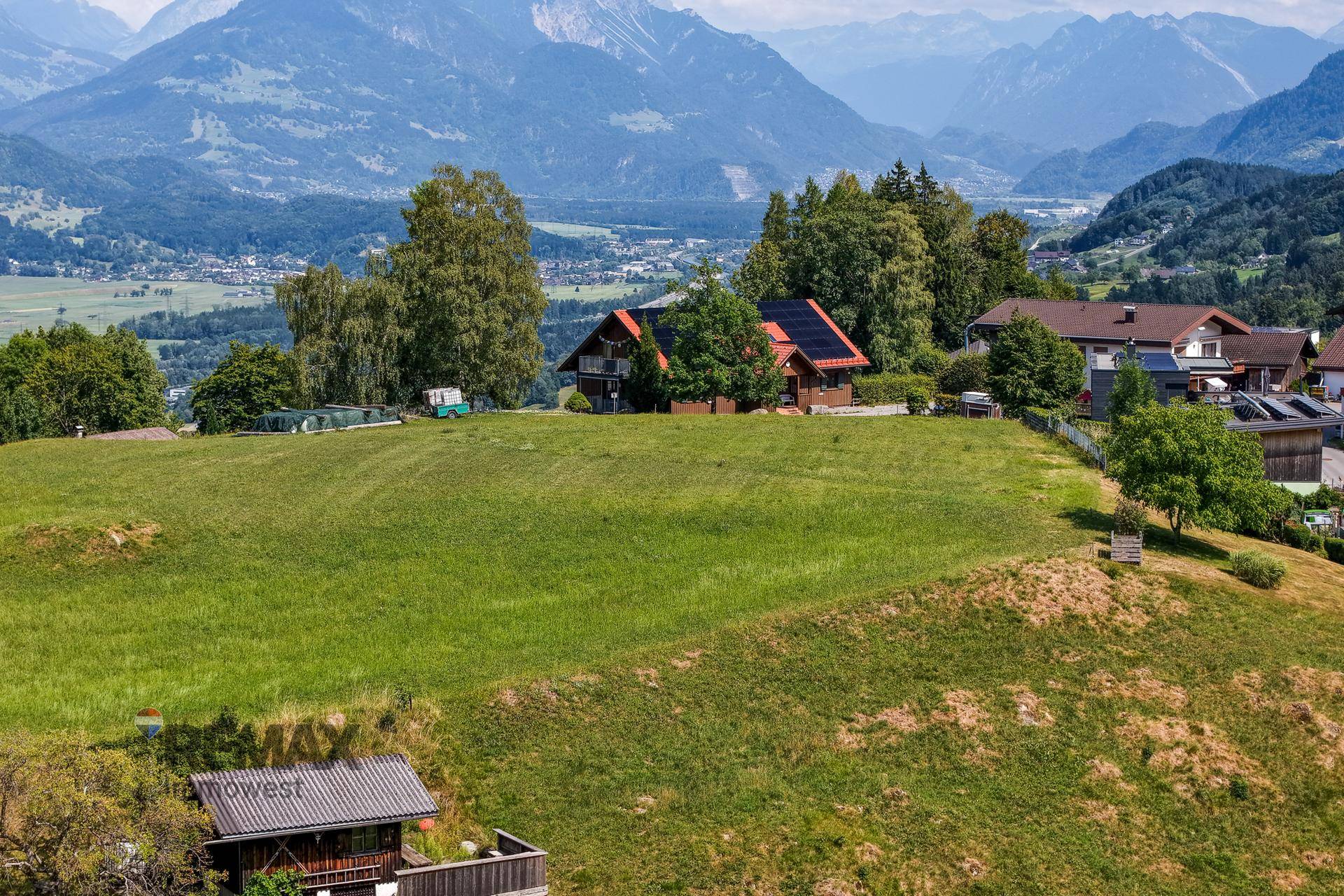 Weitläufige Wiesen und Wohngebäude mit Panoramablick auf ein Tal und die majestätischen Alpen.