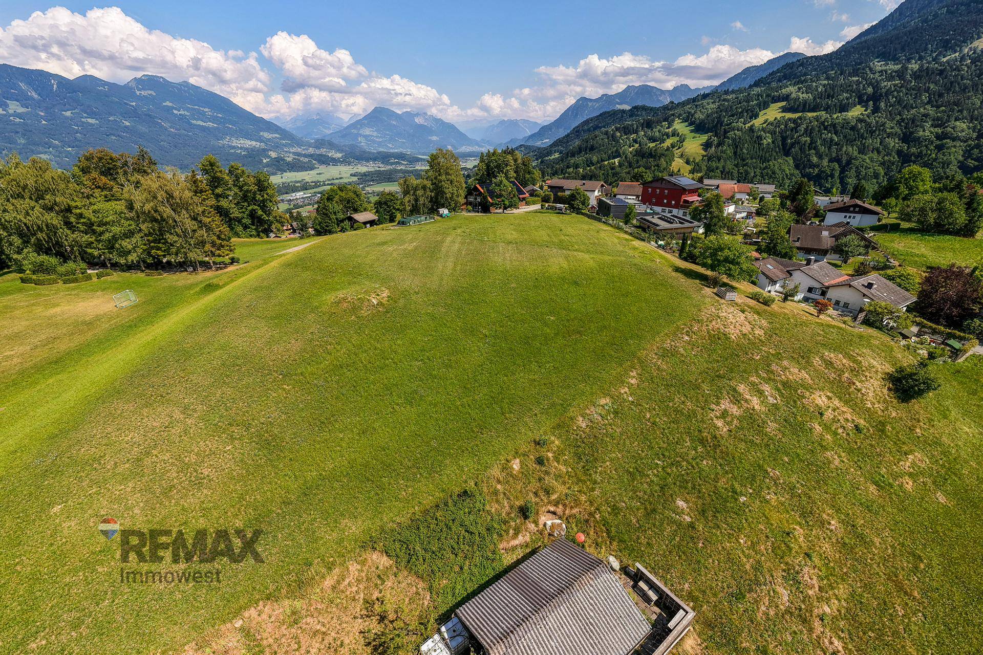 Blick über eine grüne Hanglage mit Häusern und einem Tal, umgeben von einer imposanten Bergkulisse.