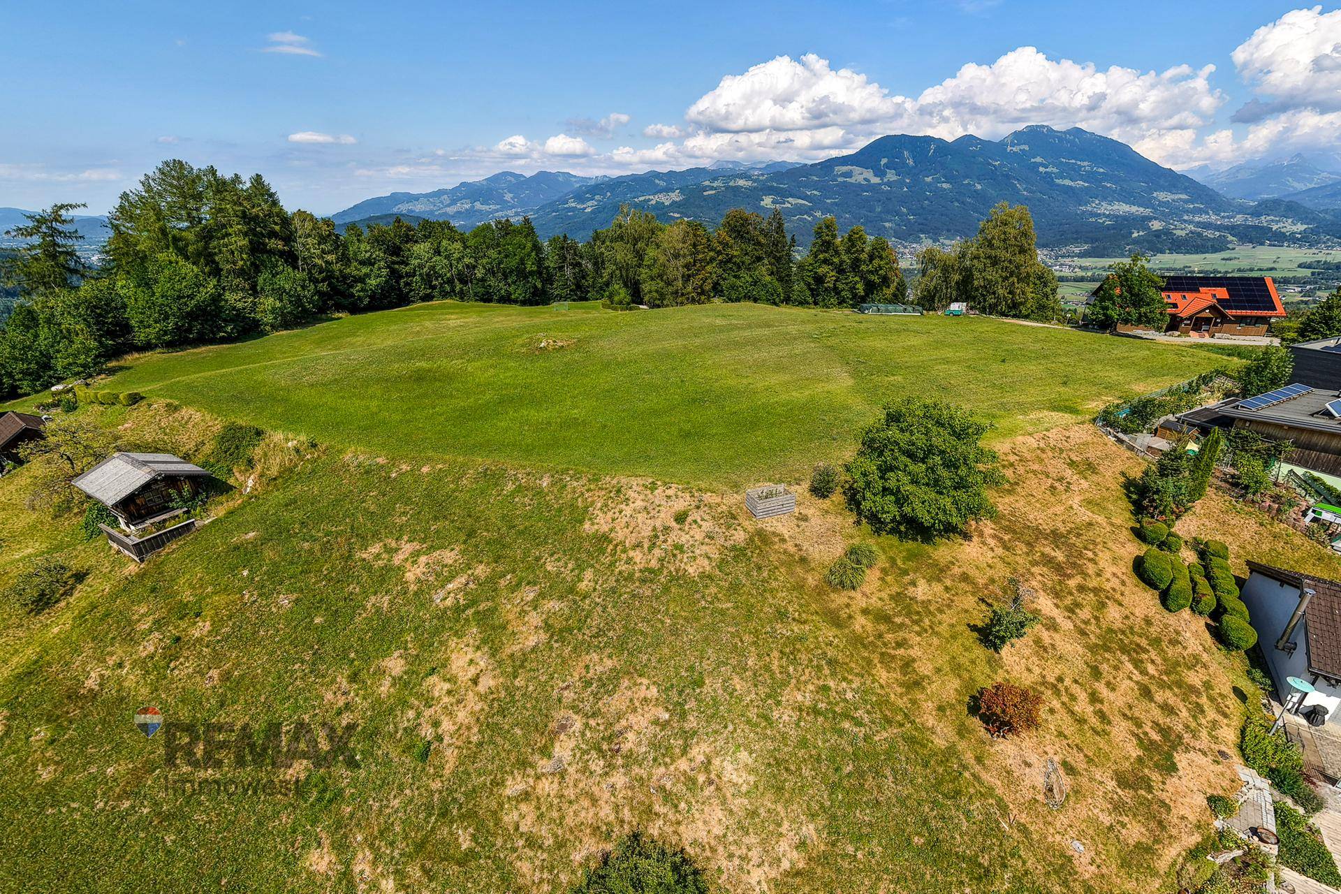 Sonniger Ausblick über grüne Wiesen und eine Siedlung mit weitem Blick auf die umliegende Bergwelt.