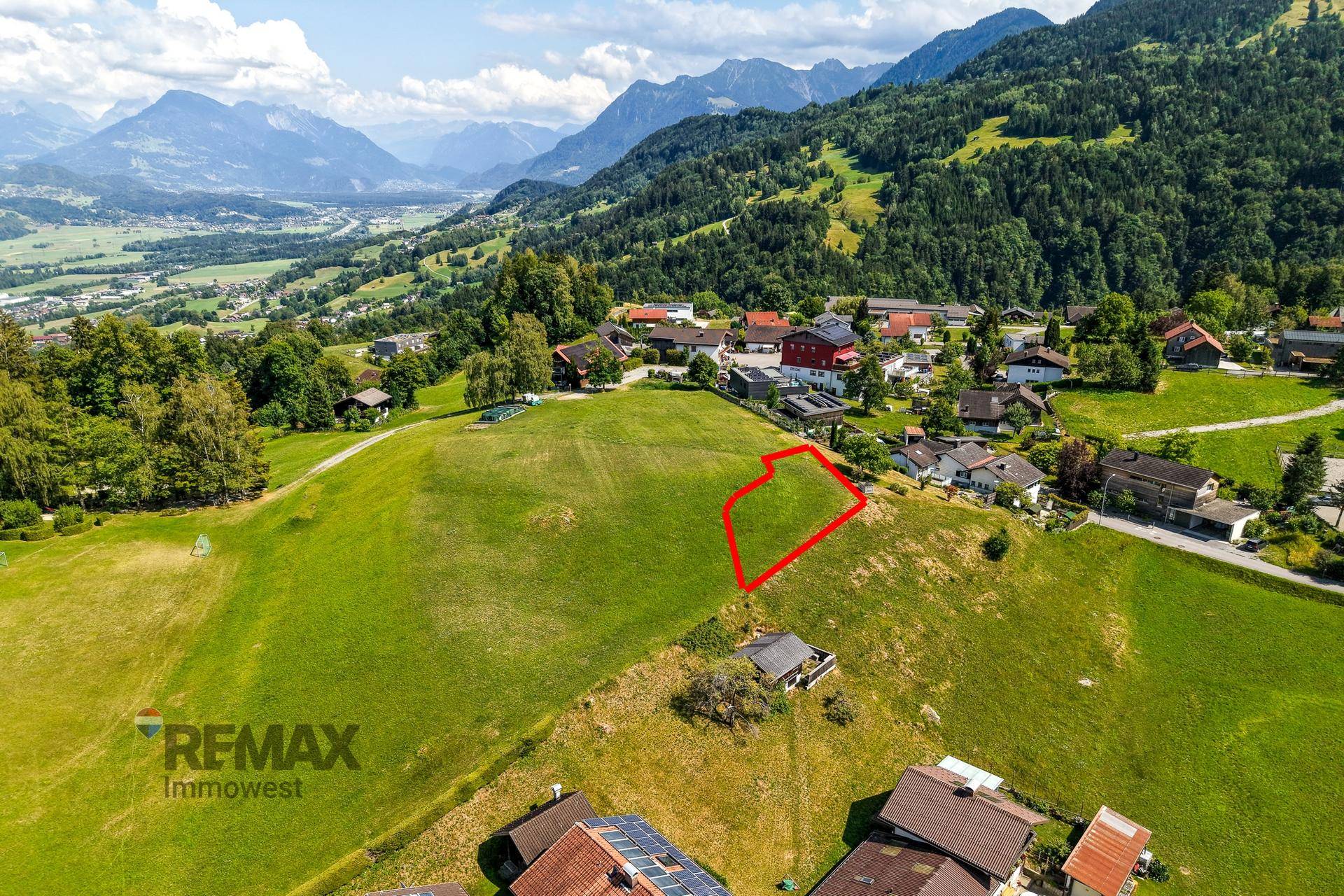 Panoramablick auf ein Baugrundstück am Hang mit roter Markierung und beeindruckendem Alpenpanorama.