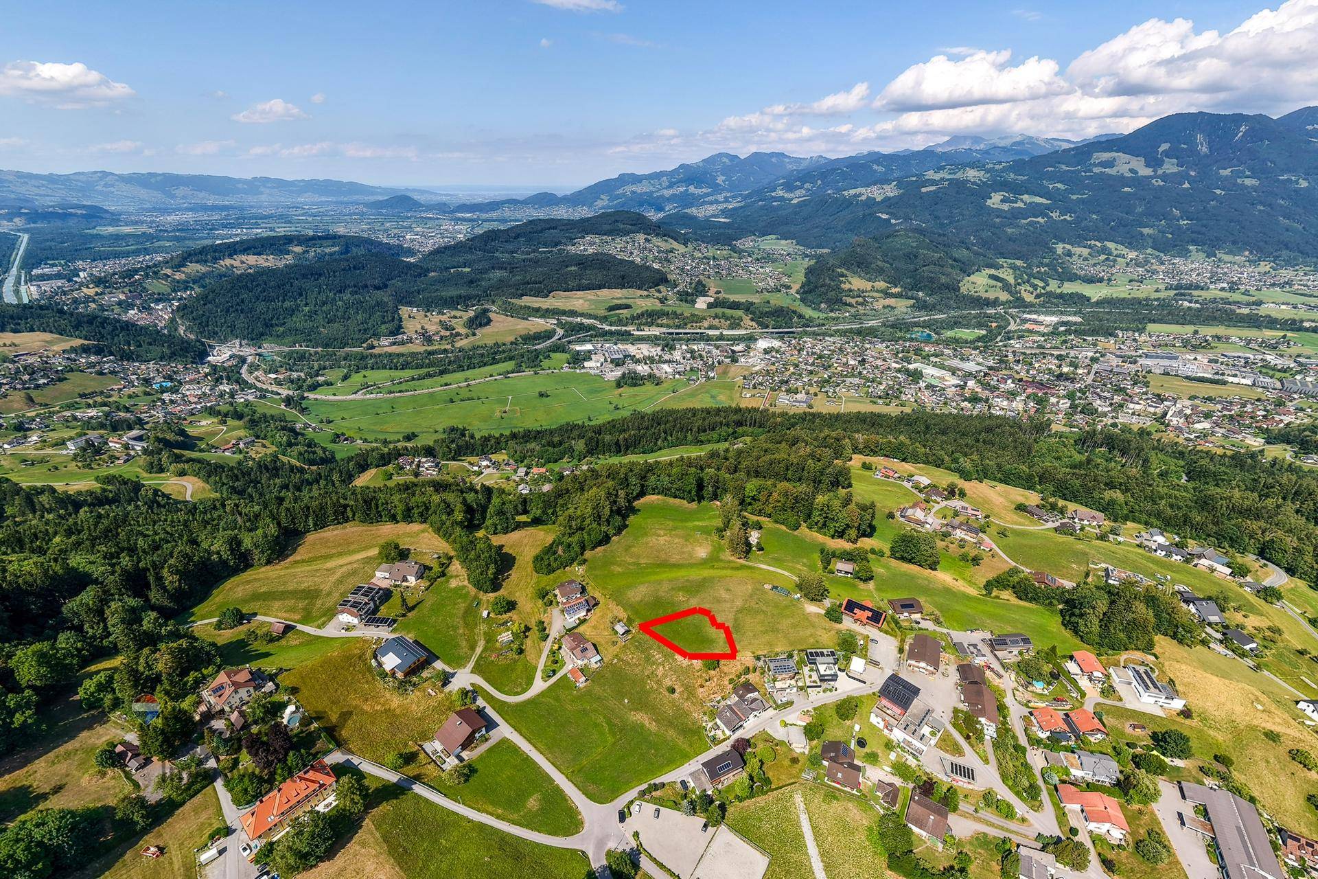 Weitwinkel-Luftaufnahme einer ländlichen Siedlung mit Baugrundstück und weitem Blick ins Tal und die Berge.