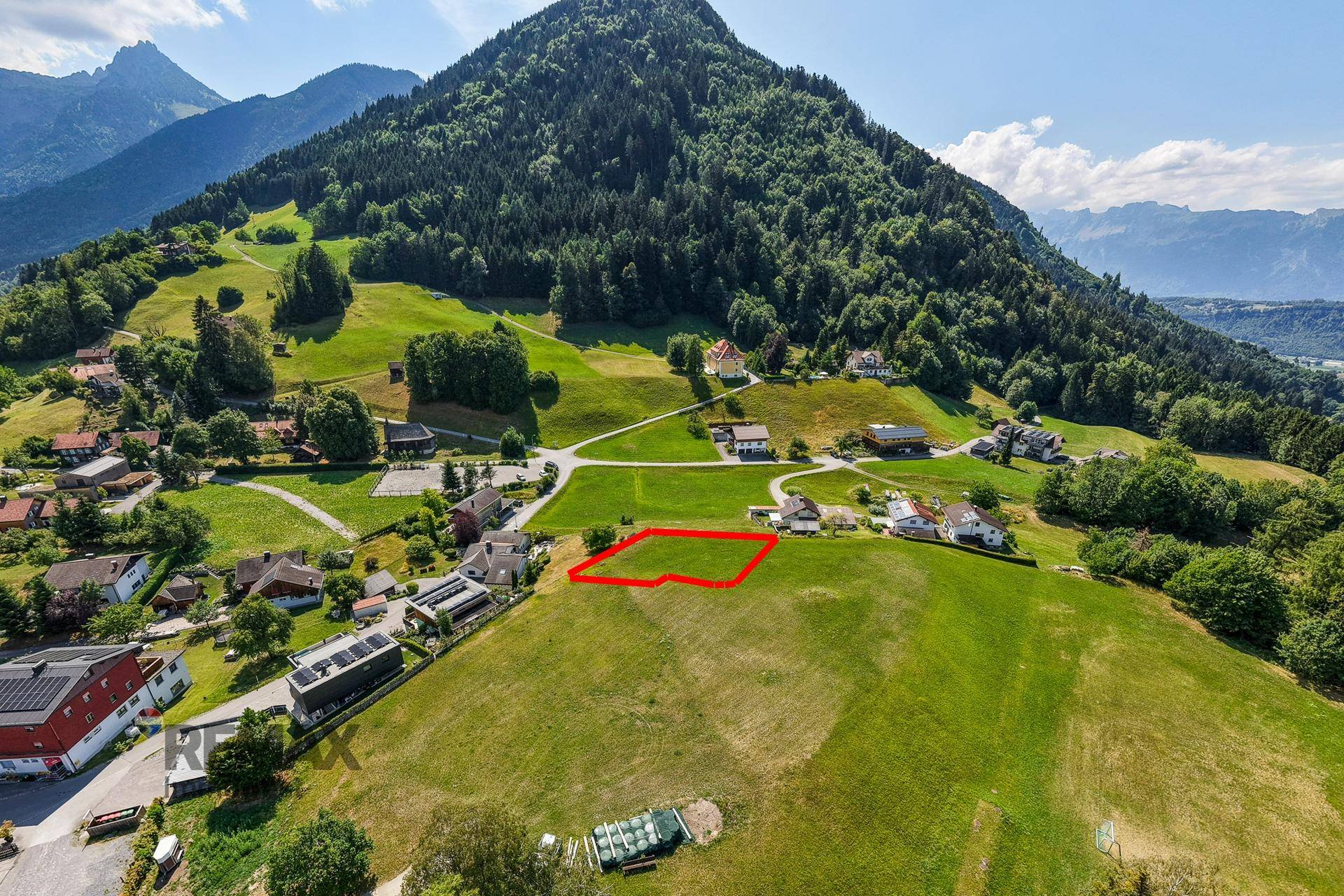 Panoramablick auf ein Baugrundstück am Hang mit roter Markierung und einem imposanten Berg im Hintergrund.