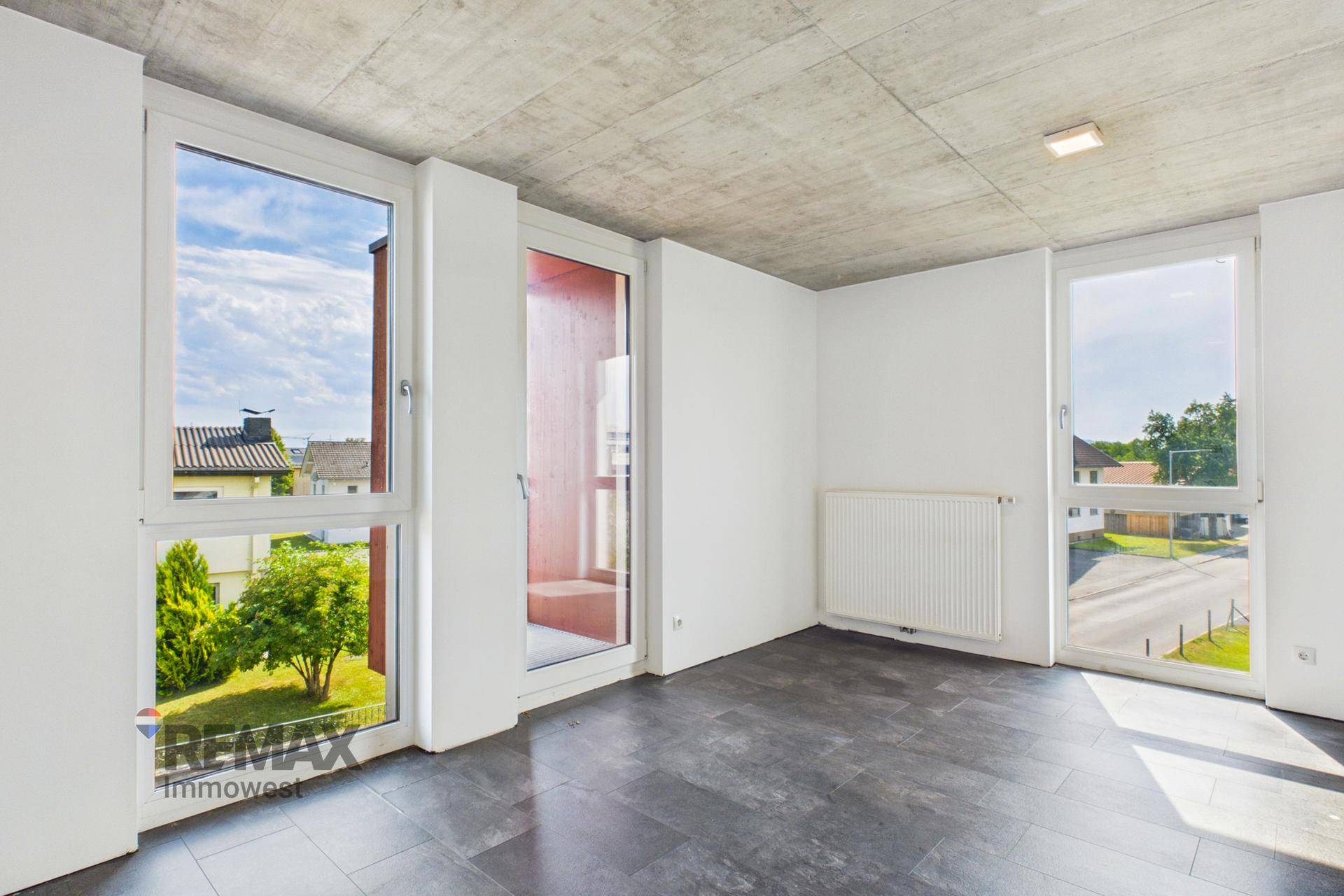 Lichtdurchfluteter Raum mit dunklen Fliesen, Betonoptikdecke und Zugang zum Balkon.