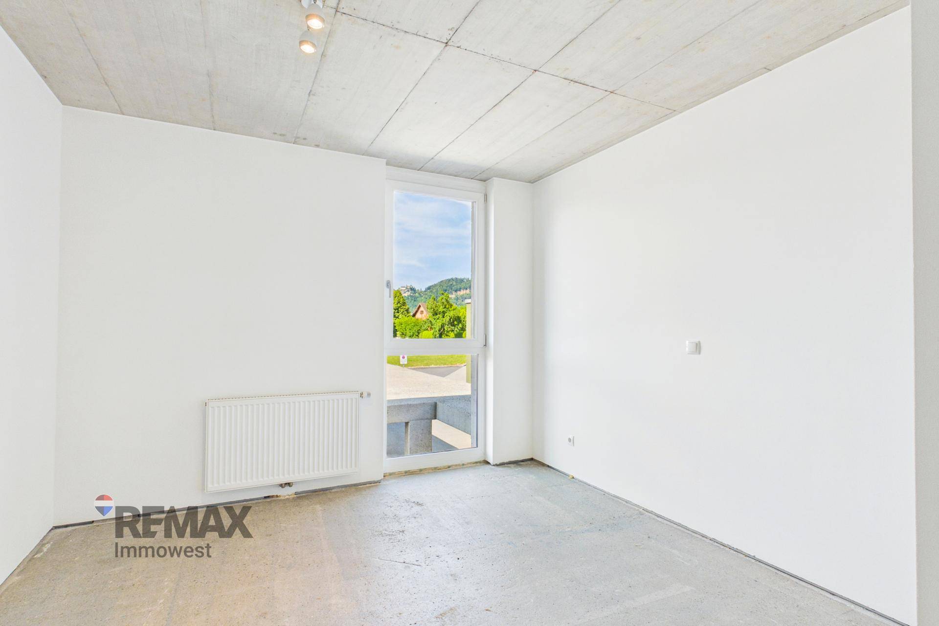 Helles Zimmer mit Betonboden, weiß gestrichenen Wänden und einem großen Fenster mit Ausblick.