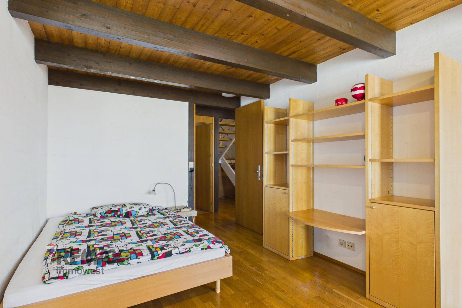 Schlafzimmer mit Einzelbett, Holzbalkendecke und integriertem Regal-Schreibtisch-System.