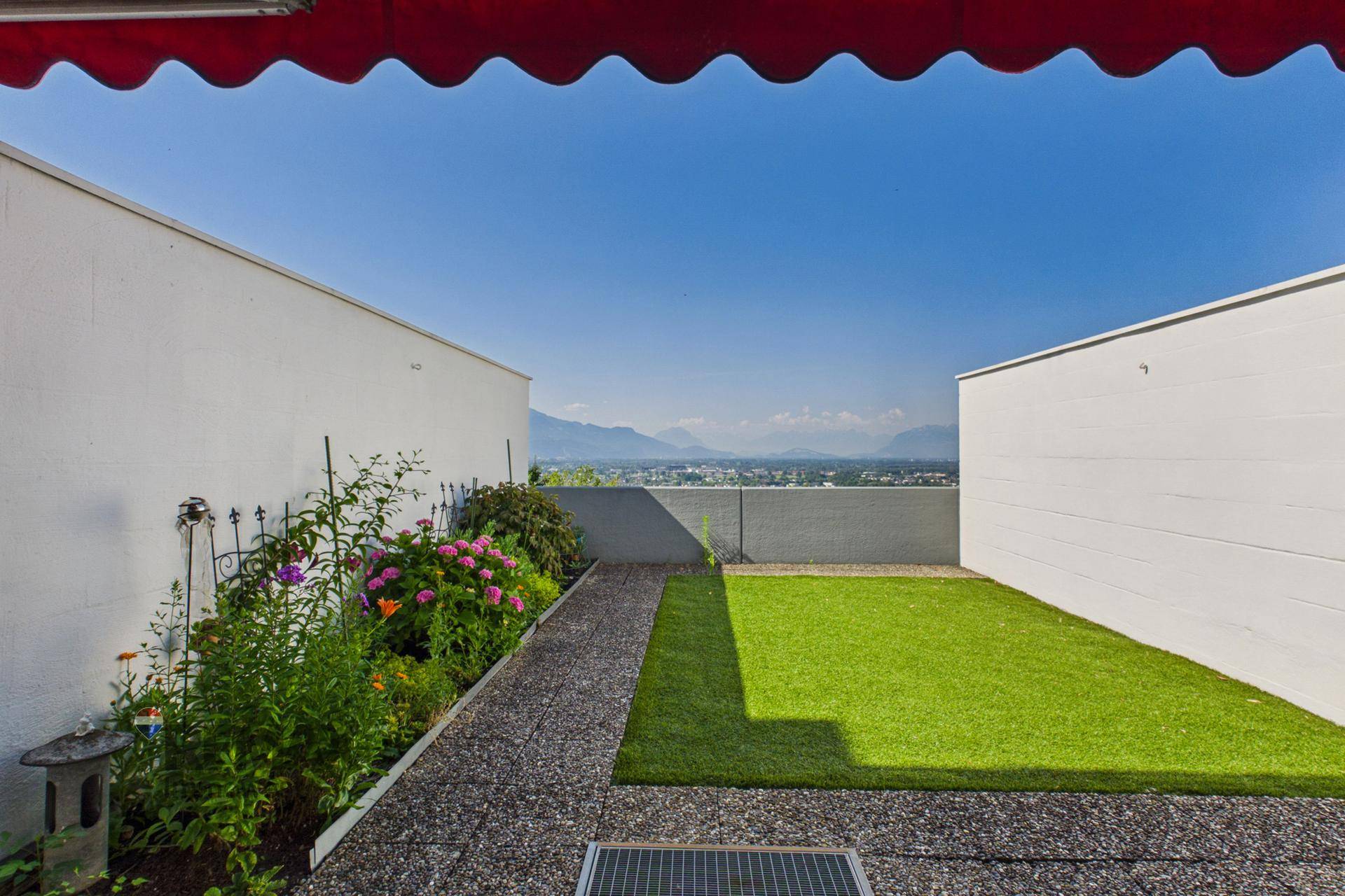 Kleiner Gartenbereich mit Kunstrasen, Blumenbeeten und Panoramablick auf die Stadt und Berge.