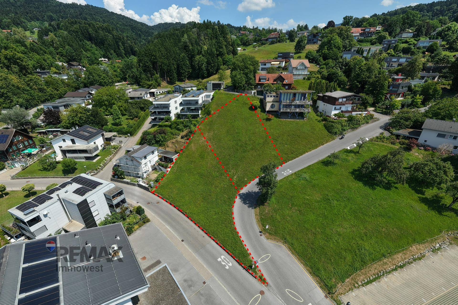 Nahaufnahme des Baugrundstücks von oben, umgeben von Wohnhäusern und einer Straße, mit Berglandschaft.