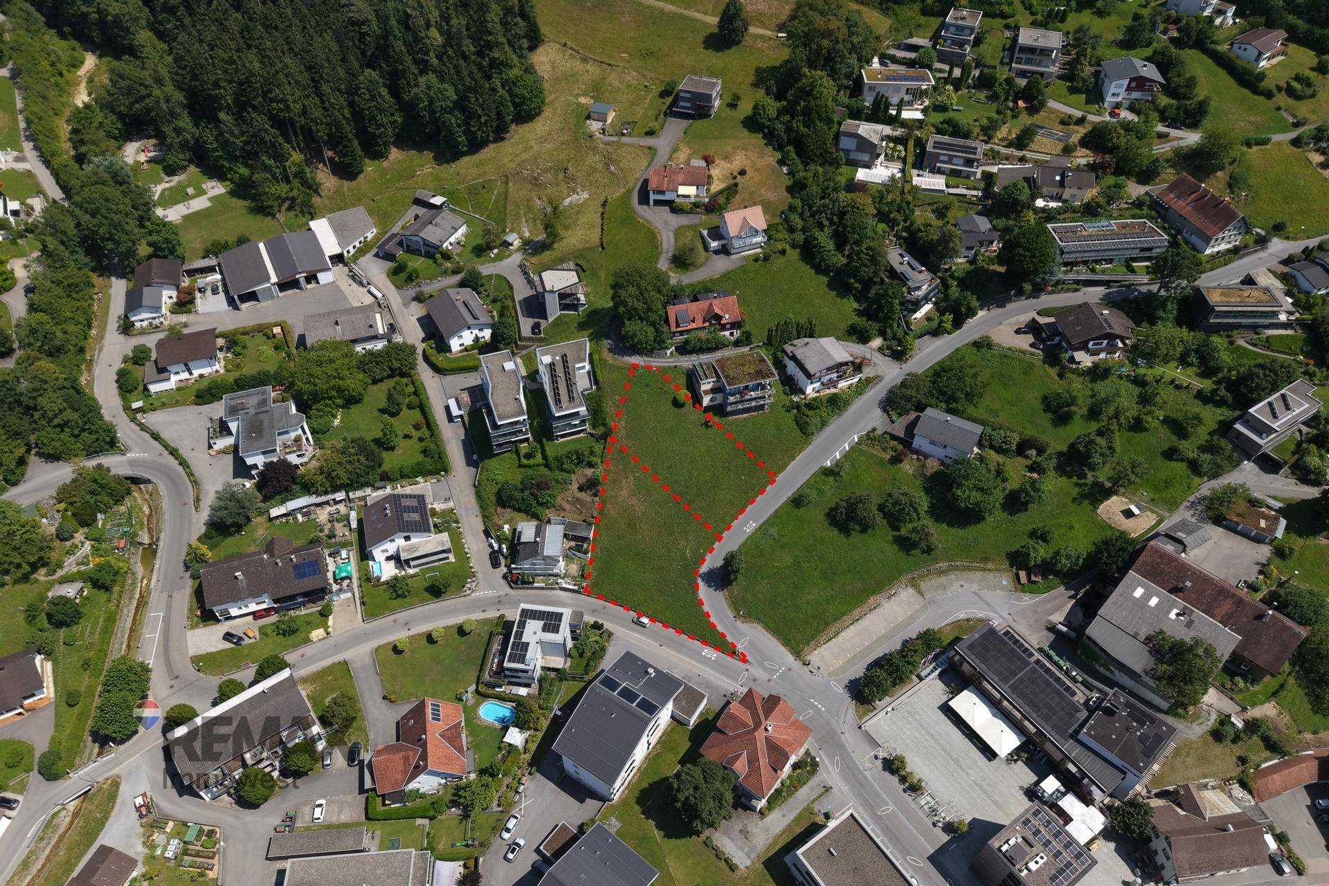 Panoramablick aus der Luft auf das Baugrundstück in einer grünen Wohngegend mit bewaldeten Bergen.