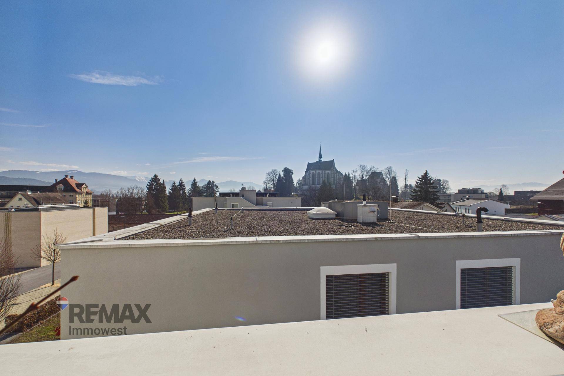 Weitblick von einem Balkon über Dächer, eine Kirche und die umliegende Berglandschaft.