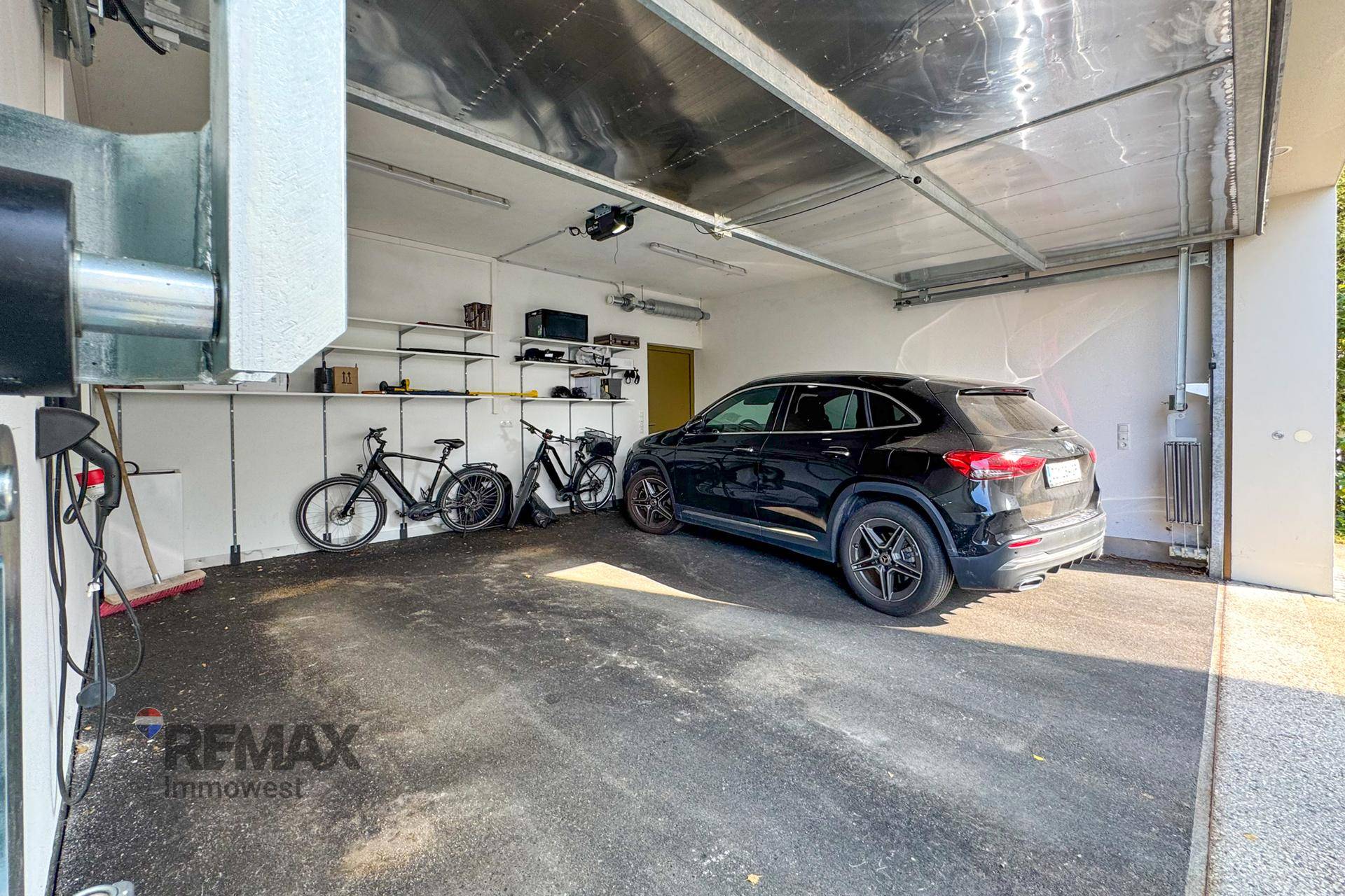 Geräumige Garage mit einem Auto, Fahrrädern, Regalen und einer Ladestation für Elektrofahrzeuge.