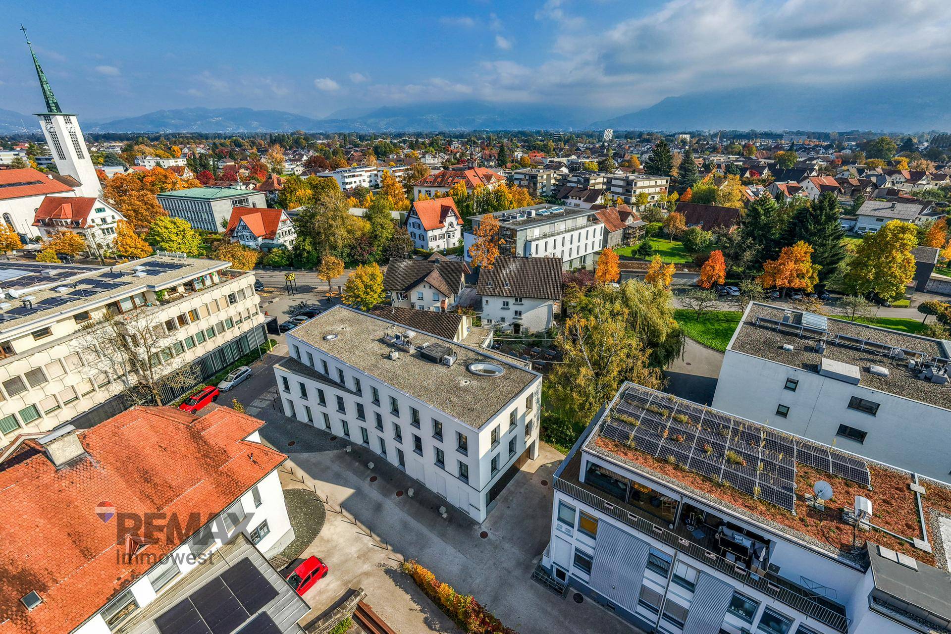 Weitläufige Luftaufnahme einer Stadt mit Kirche, Wohngebäuden und einer beeindruckenden Bergkulisse.