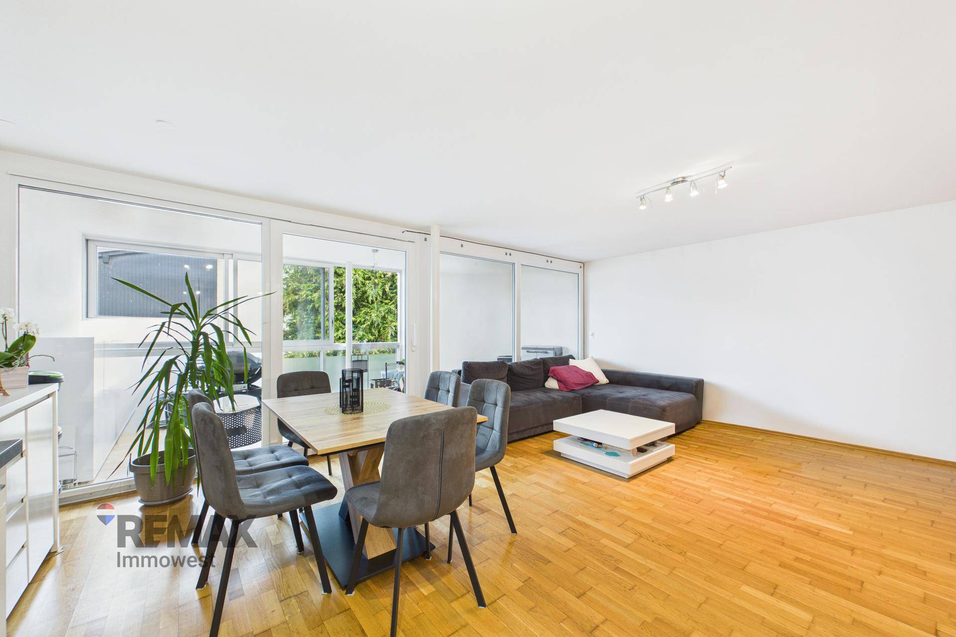 Offener Wohn- und Essbereich mit Holzboden und großen Fenstern zum Balkon.