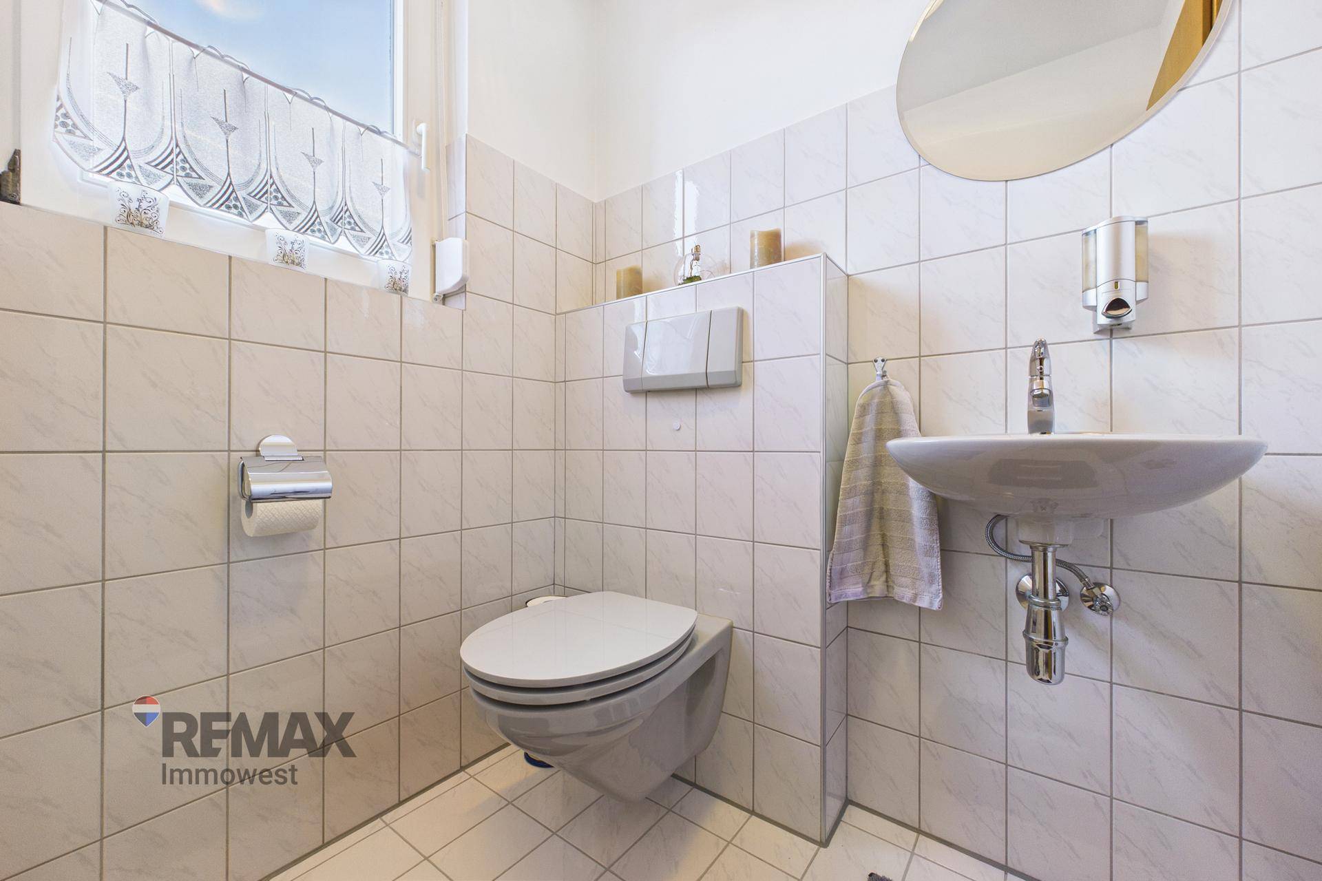 Helles Gäste-WC mit Wand-WC, Waschbecken und Fenster in neutralen Fliesen.