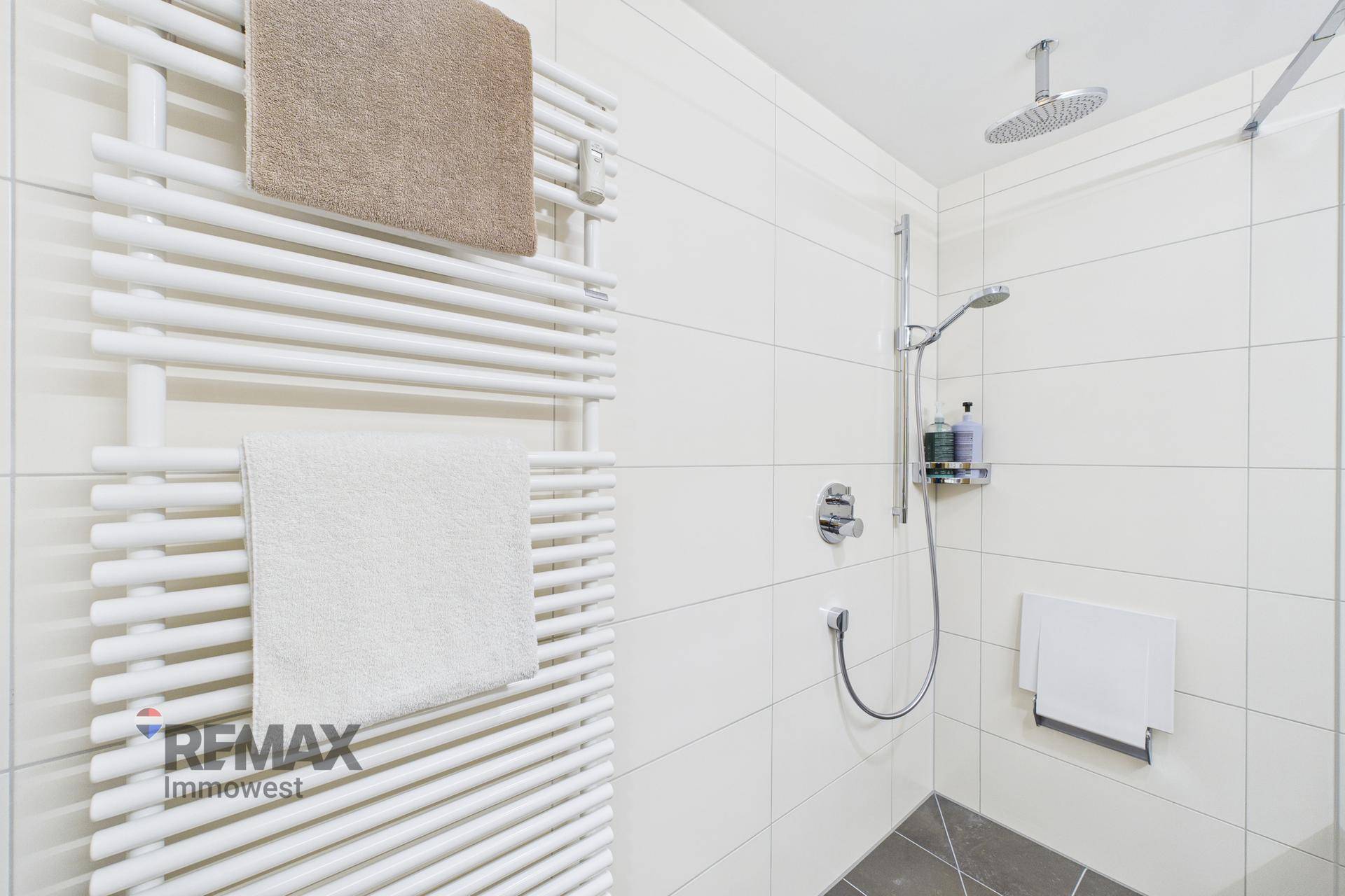 Moderne Dusche mit Regenduschkopf und beheiztem Handtuchhalter in weiß gefliestem Bad.