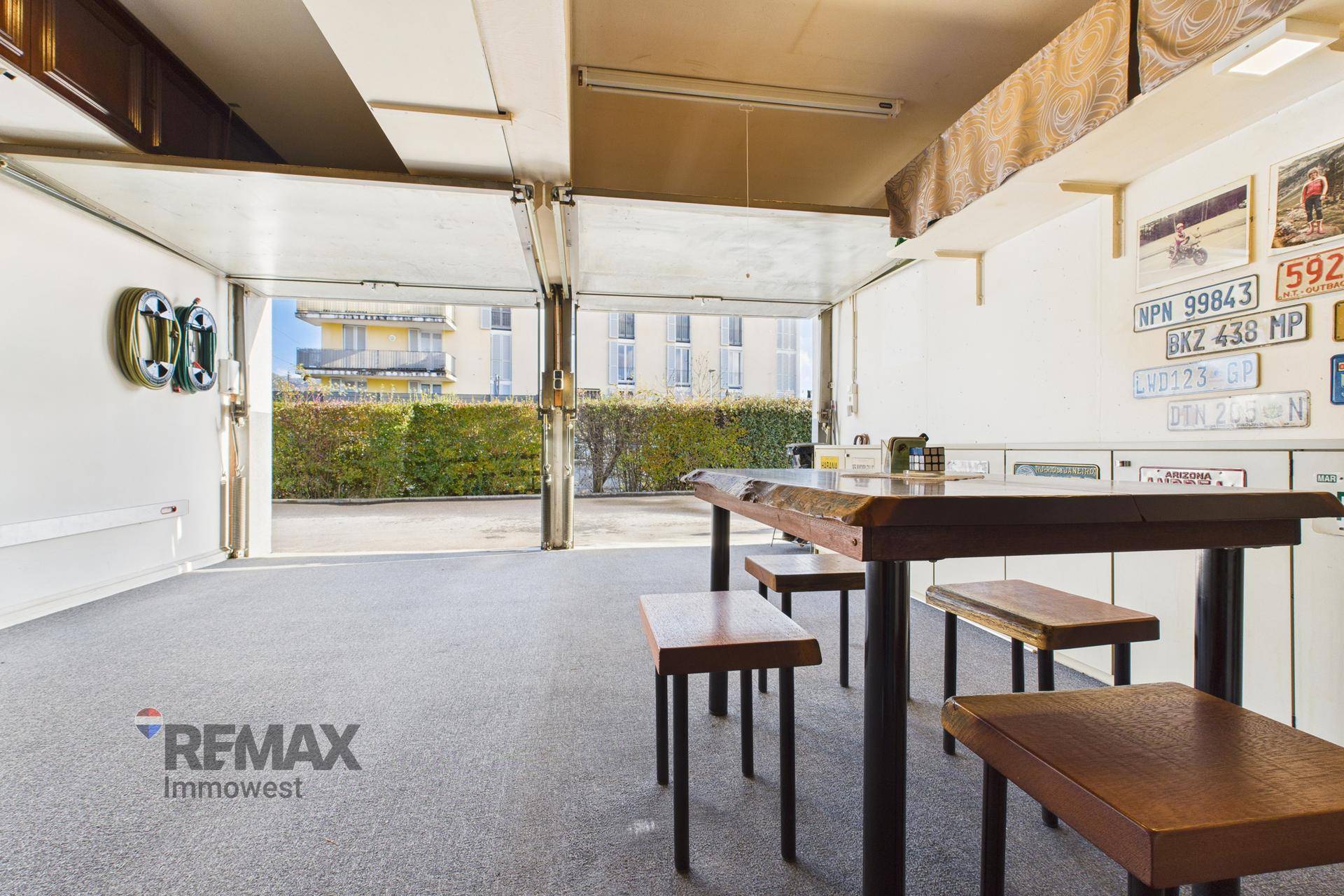 Garage mit Teppichboden, Tisch und Hockern, offen zur Einfahrt und Außenbereich.