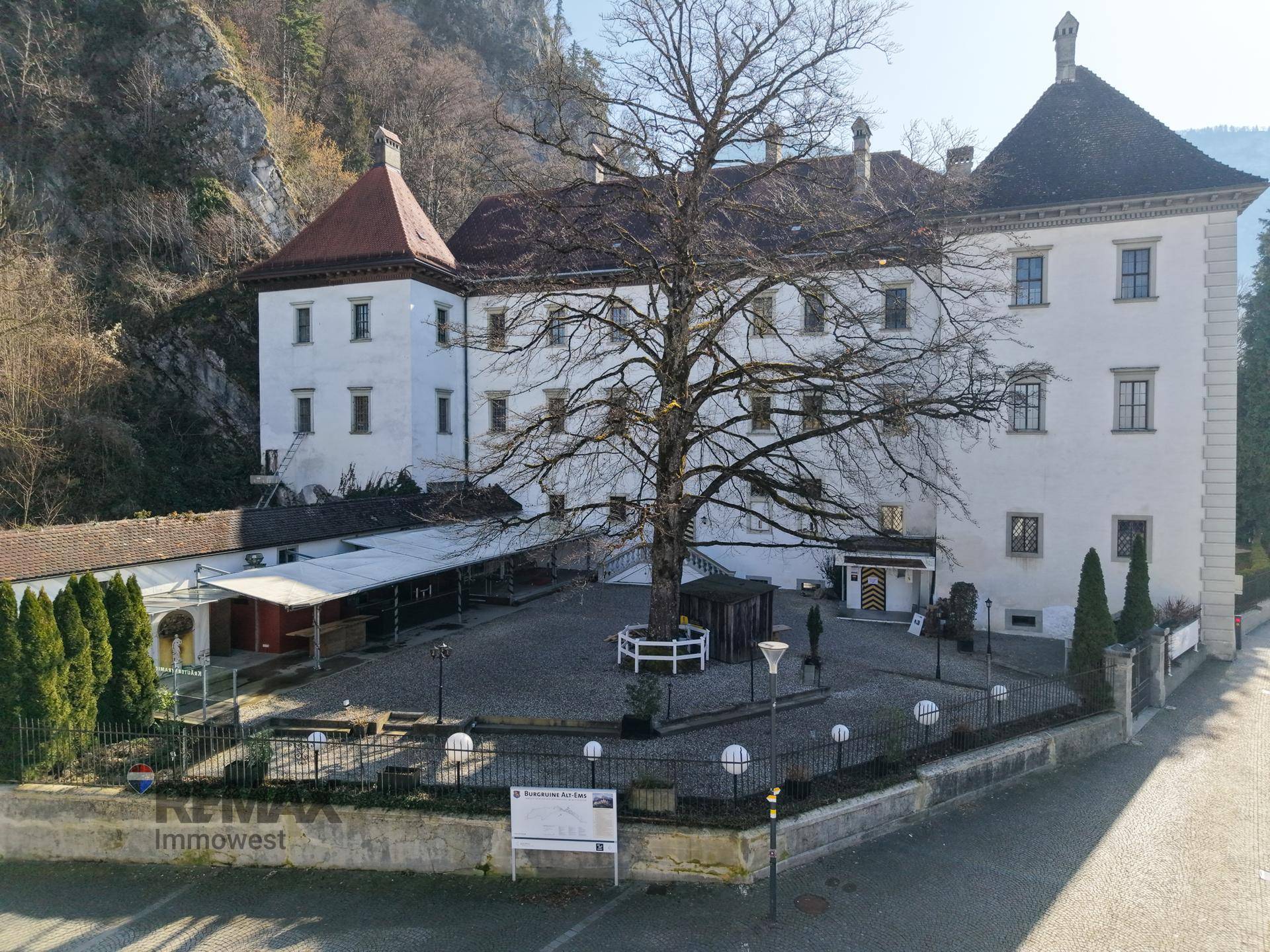 Historisches Gebäude mit großem Innenhof und altem Baum, umgeben von natürlicher Landschaft.