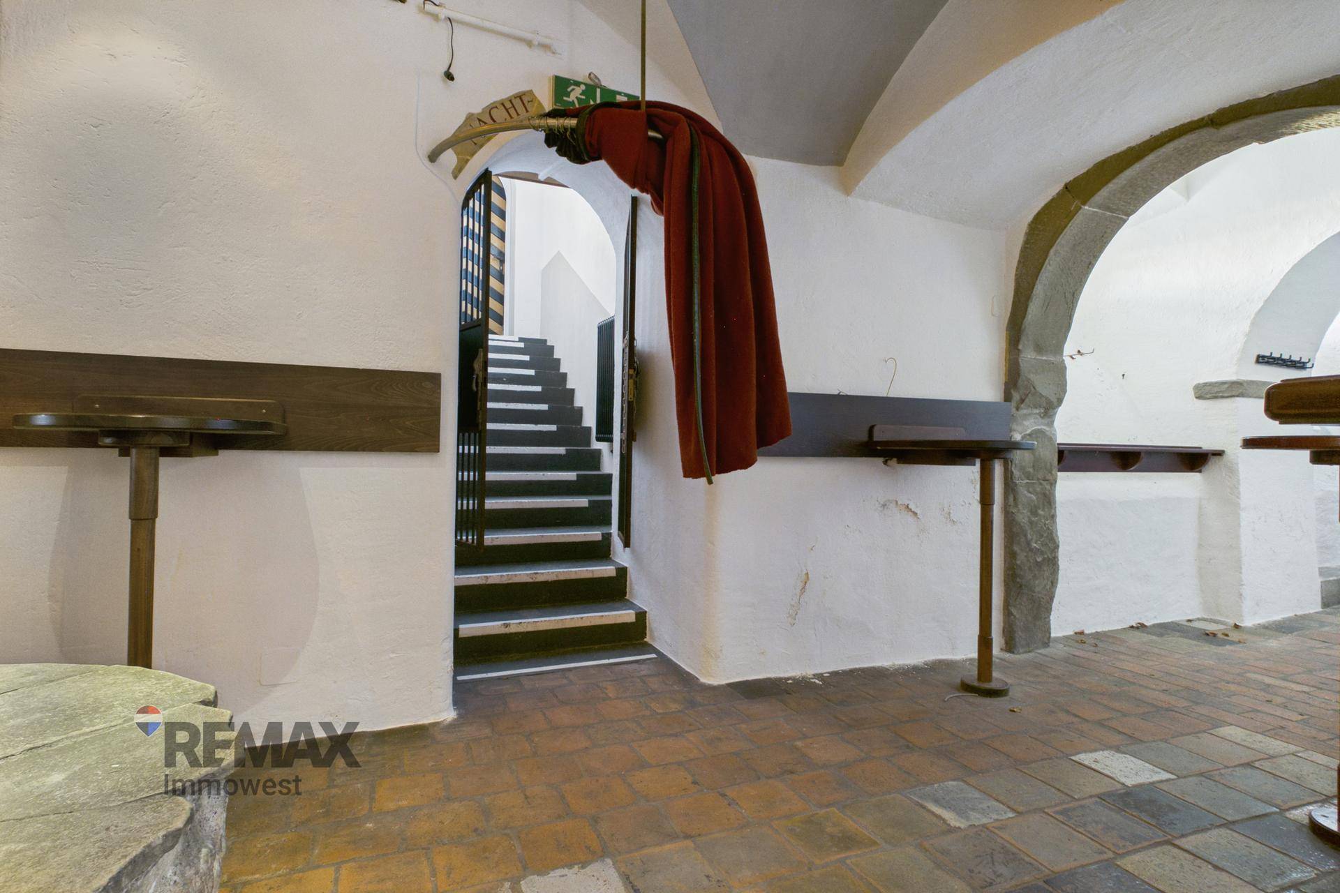 Historischer Flur mit Steintischen, Ziegelboden und einem Treppenaufgang im Hintergrund.