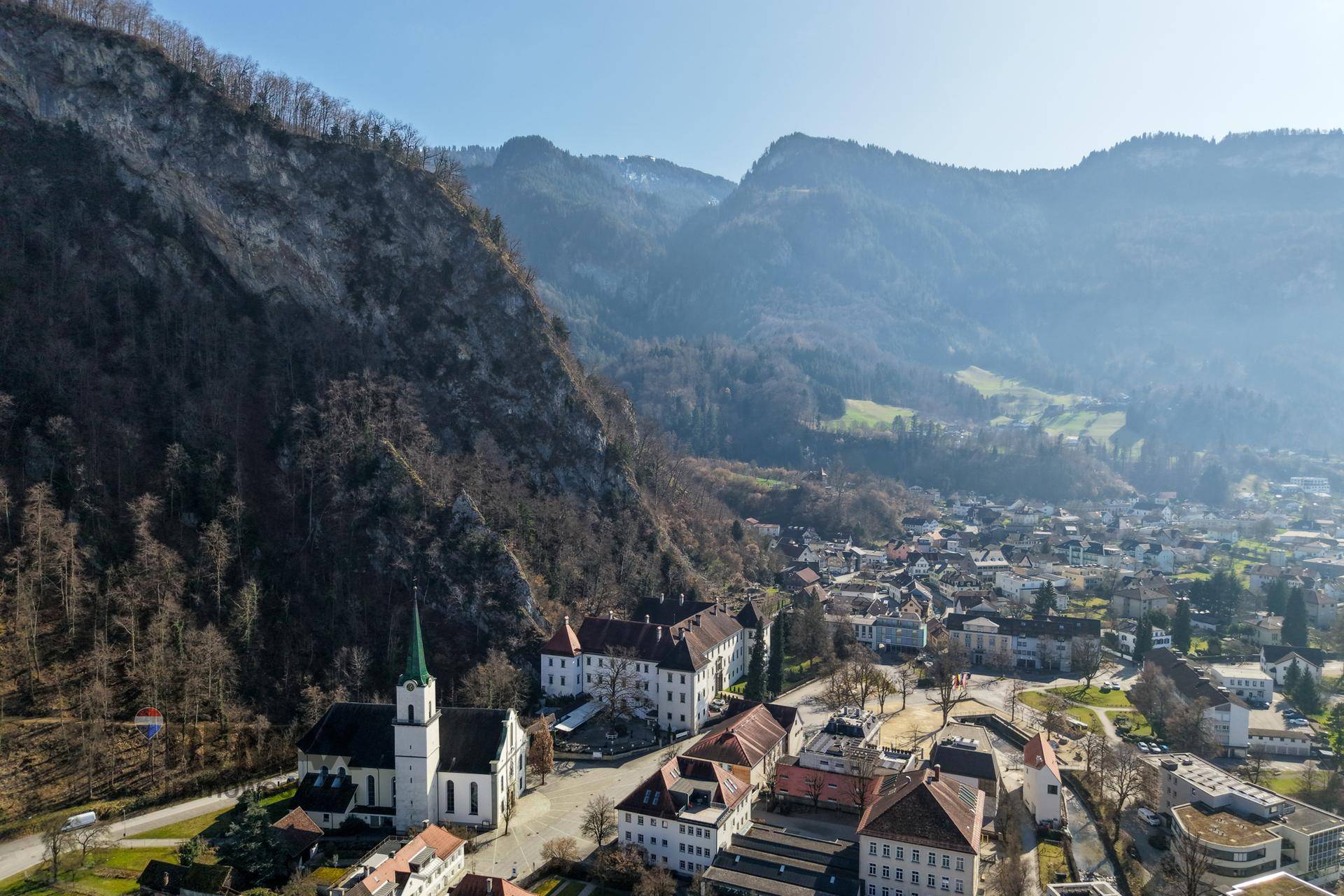 Luftaufnahme einer historischen Stadt mit Kirche und Klosteranlage, umgeben von majestätischen Bergen und grünen Tälern.