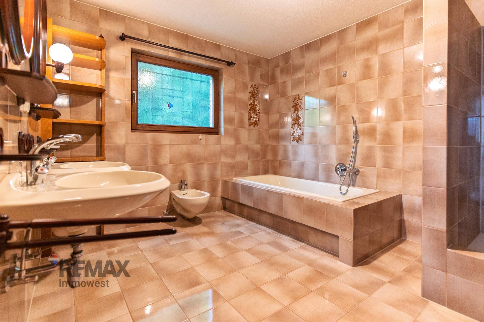 Gefliestes Badezimmer in warmen Brauntönen mit Doppelwaschbecken, Badewanne und Fenster.