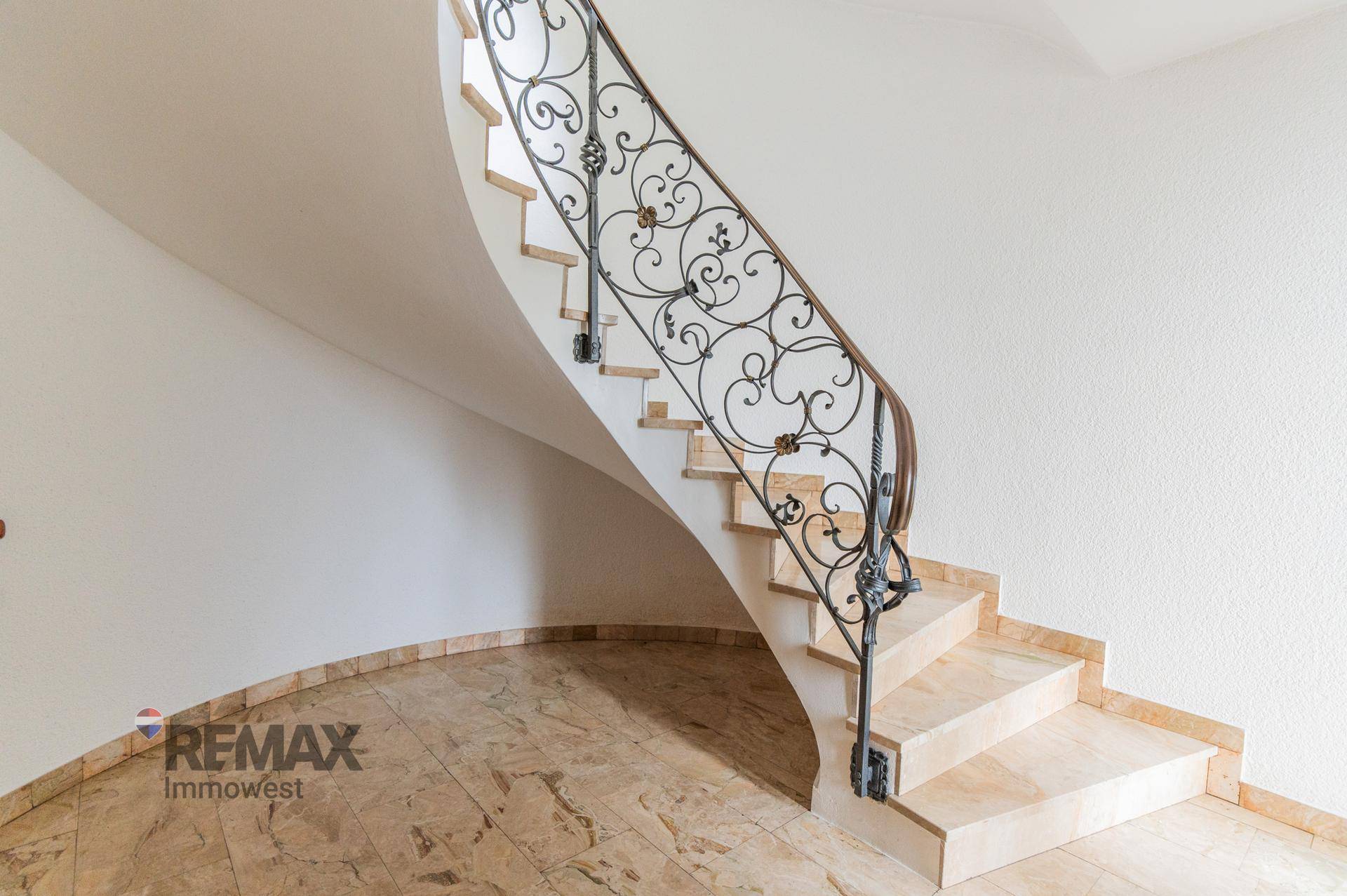 Elegante geschwungene Treppe mit schmiedeeisernem Geländer und Marmorboden in einem hellen Flur.