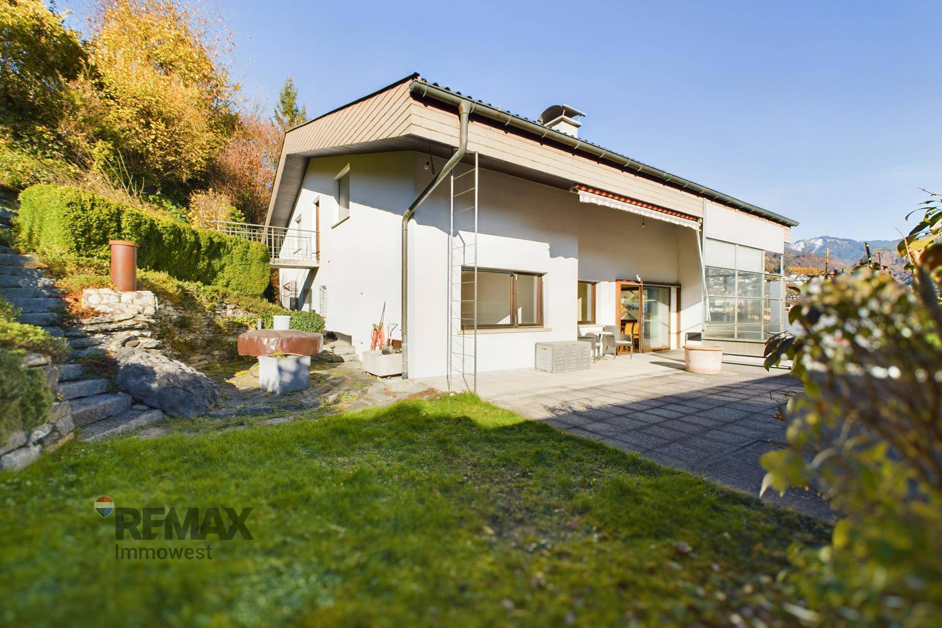 Hausfassade mit Terrasse und gestuftem Gartenbereich, umgeben von grüner Natur und Bergen.