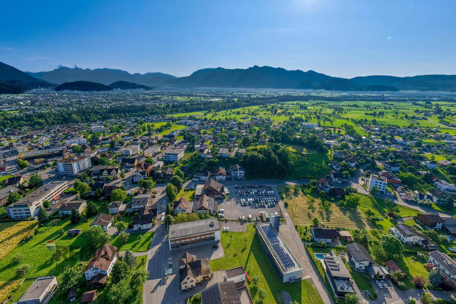 Panoramische Luftaufnahme einer weitläufigen Stadtlandschaft mit grünen Feldern und Bergen unter blauem Himmel.