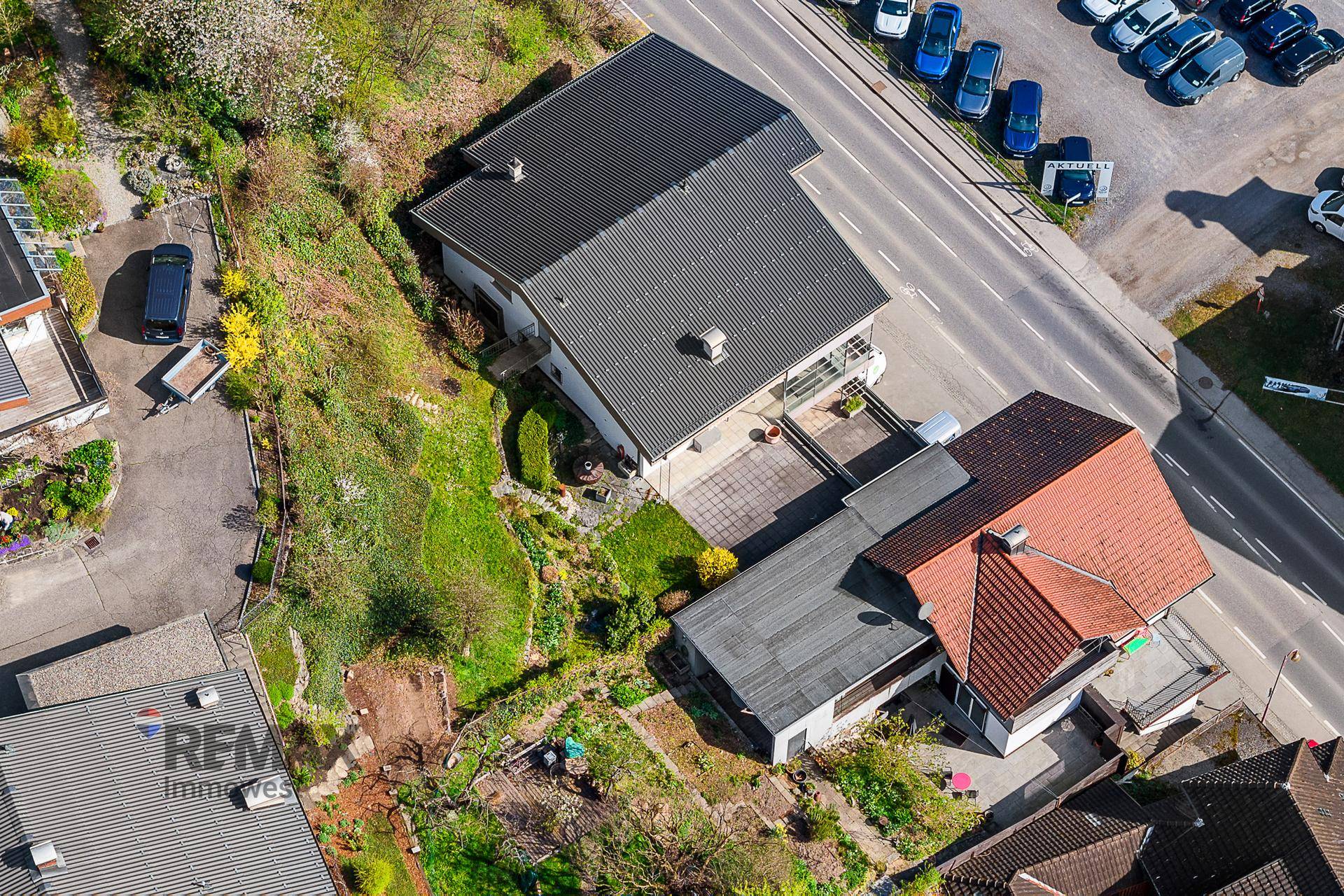 Luftaufnahme eines Wohnhauses mit dunklem Dach, angrenzendem Gebäude, Garten und einer Straße mit parkenden Autos.