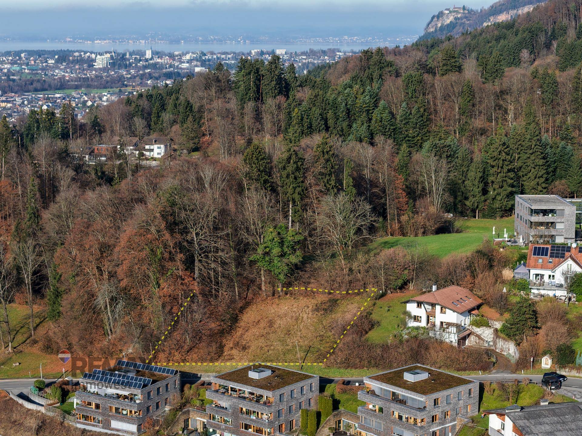 Luftaufnahme eines Grundstücks in Hanglage, umgeben von Wald und Wohnhäusern mit Fernblick.