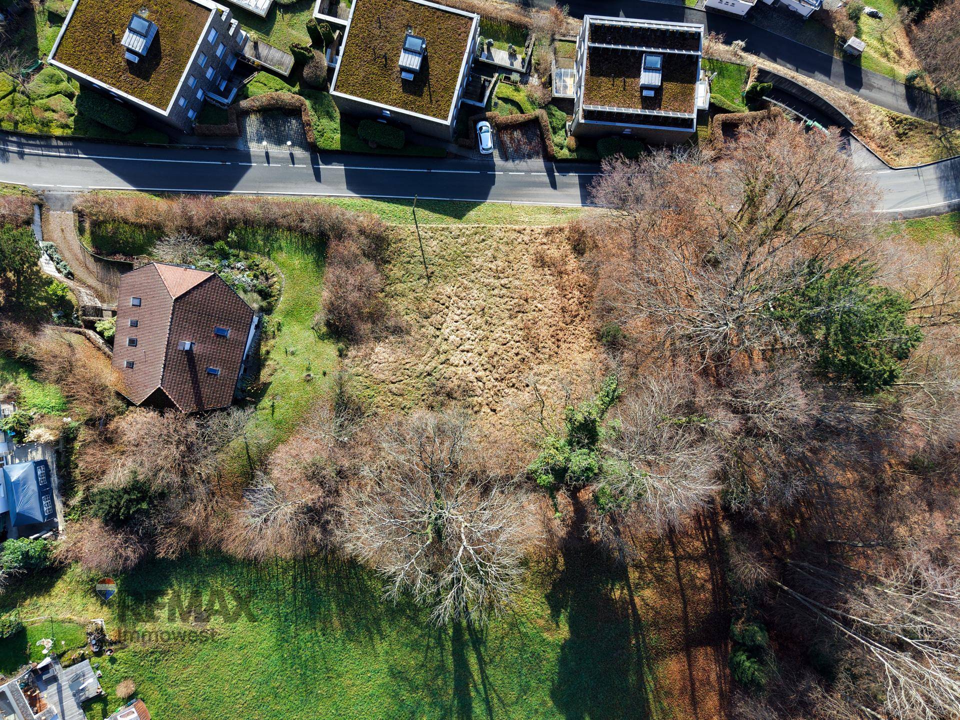 Senkrechte Luftaufnahme eines unbebauten Grundstücks neben einer Straße, umgeben von Bäumen und einem Haus.