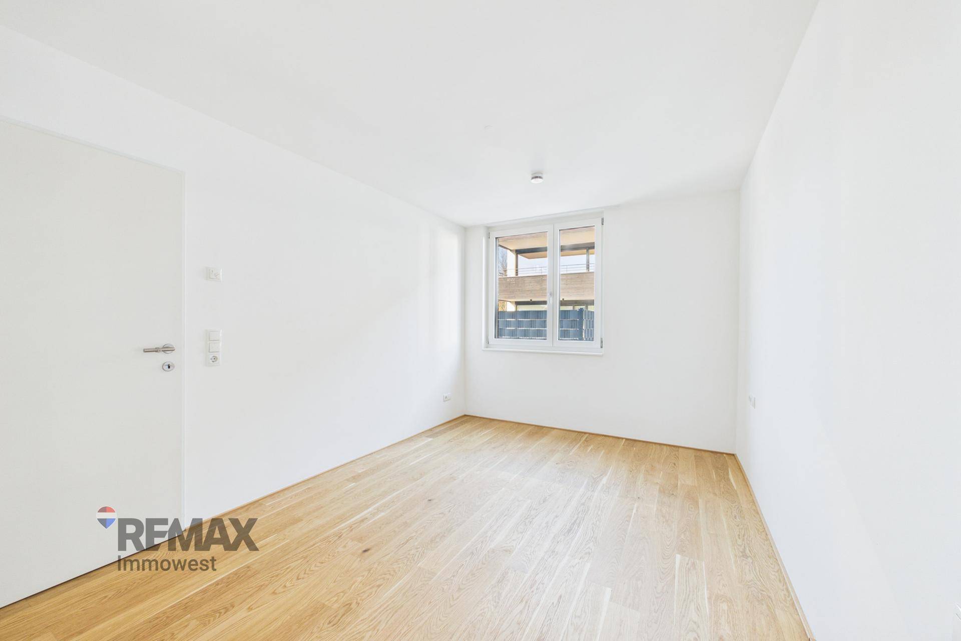 Helles, leeres Zimmer mit Holzboden und einem Fenster, das den Blick nach draußen freigibt.