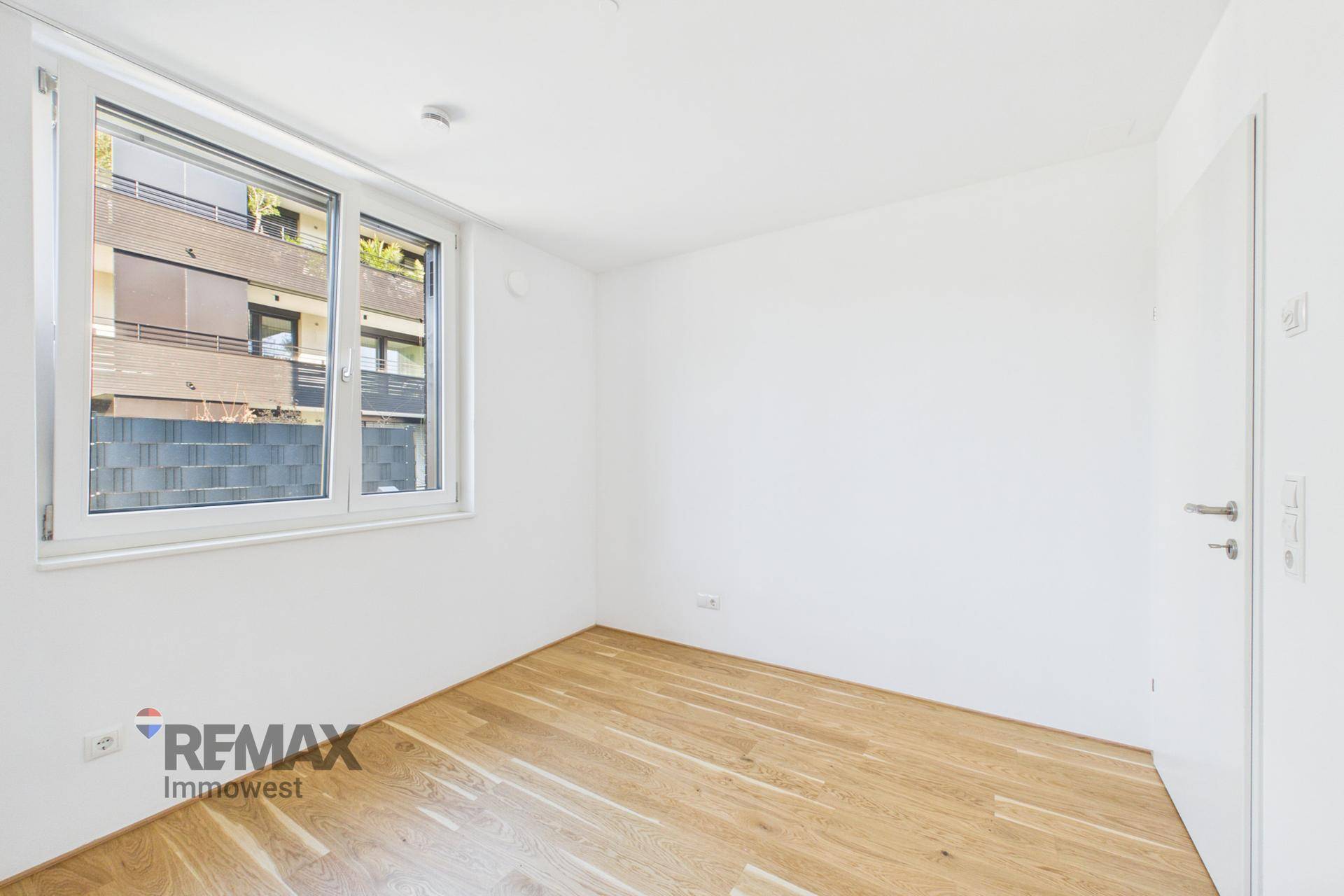 Leeres Zimmer mit Holzboden und einem Fenster, das den Blick auf die Umgebung bietet.
