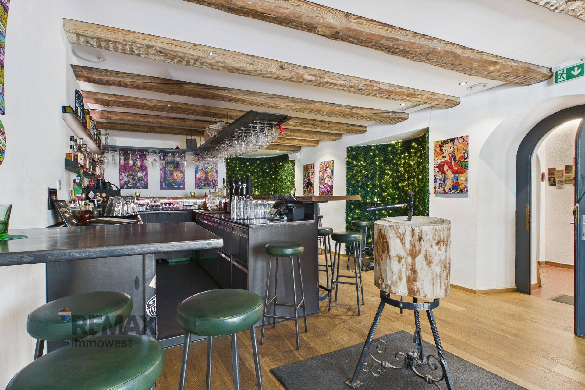 Bar mit langer Theke, Barhockern, rustikalen Holzbalken und dekorativen grünen Wänden mit Lichtern.