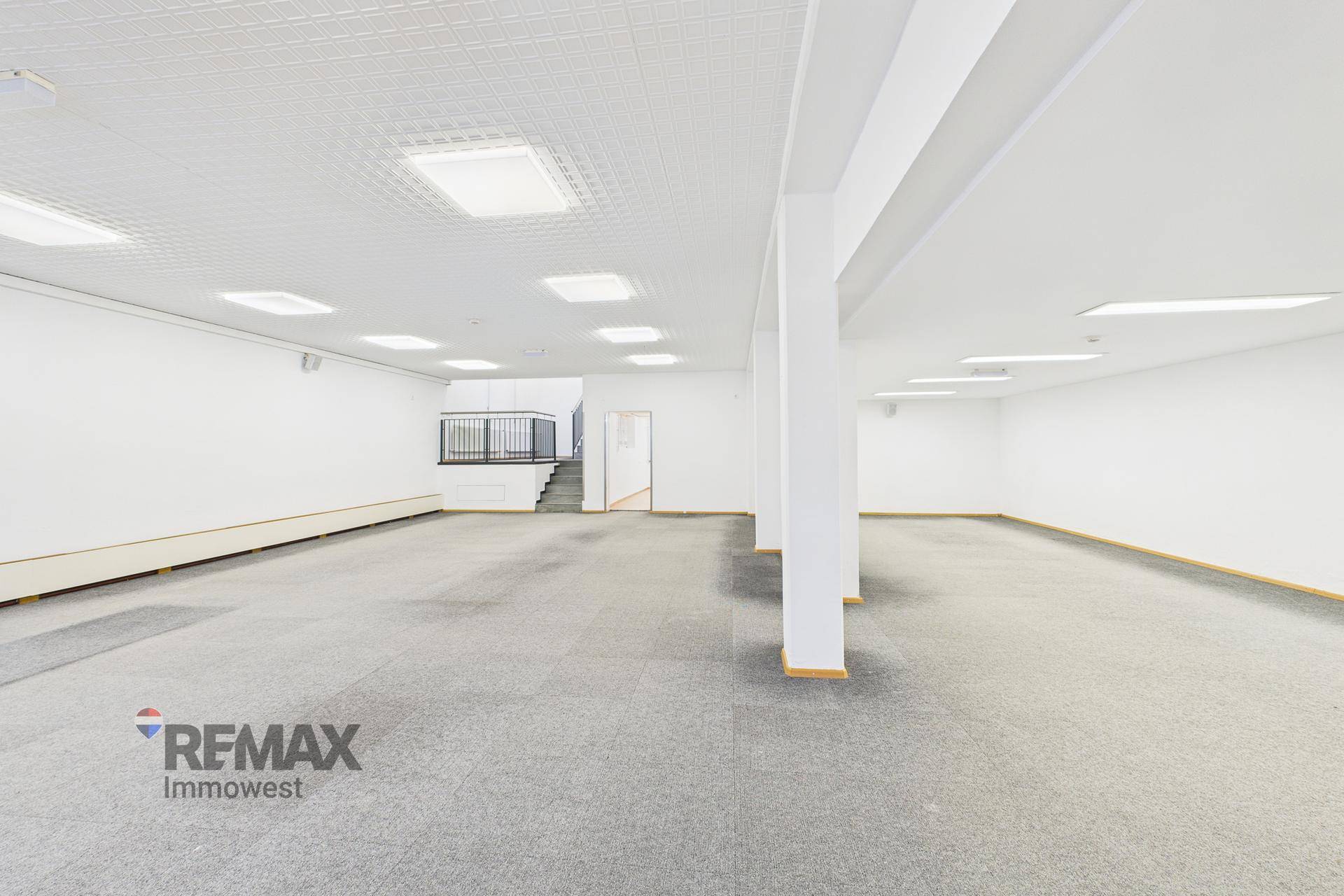 Großer, offener Gewerberaum mit Säulen, grauem Teppichboden und vielen Deckenleuchten.