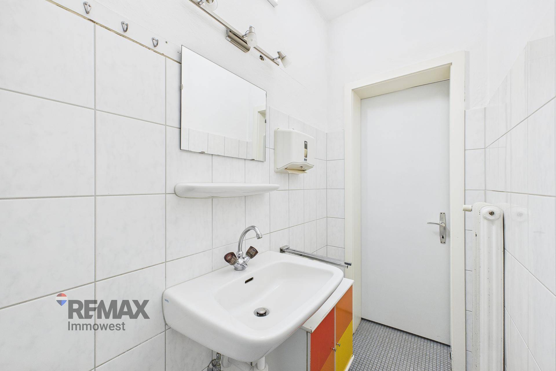 Kleines Badezimmer mit weißen Fliesen, Waschbecken und Spiegel, funktional eingerichtet.