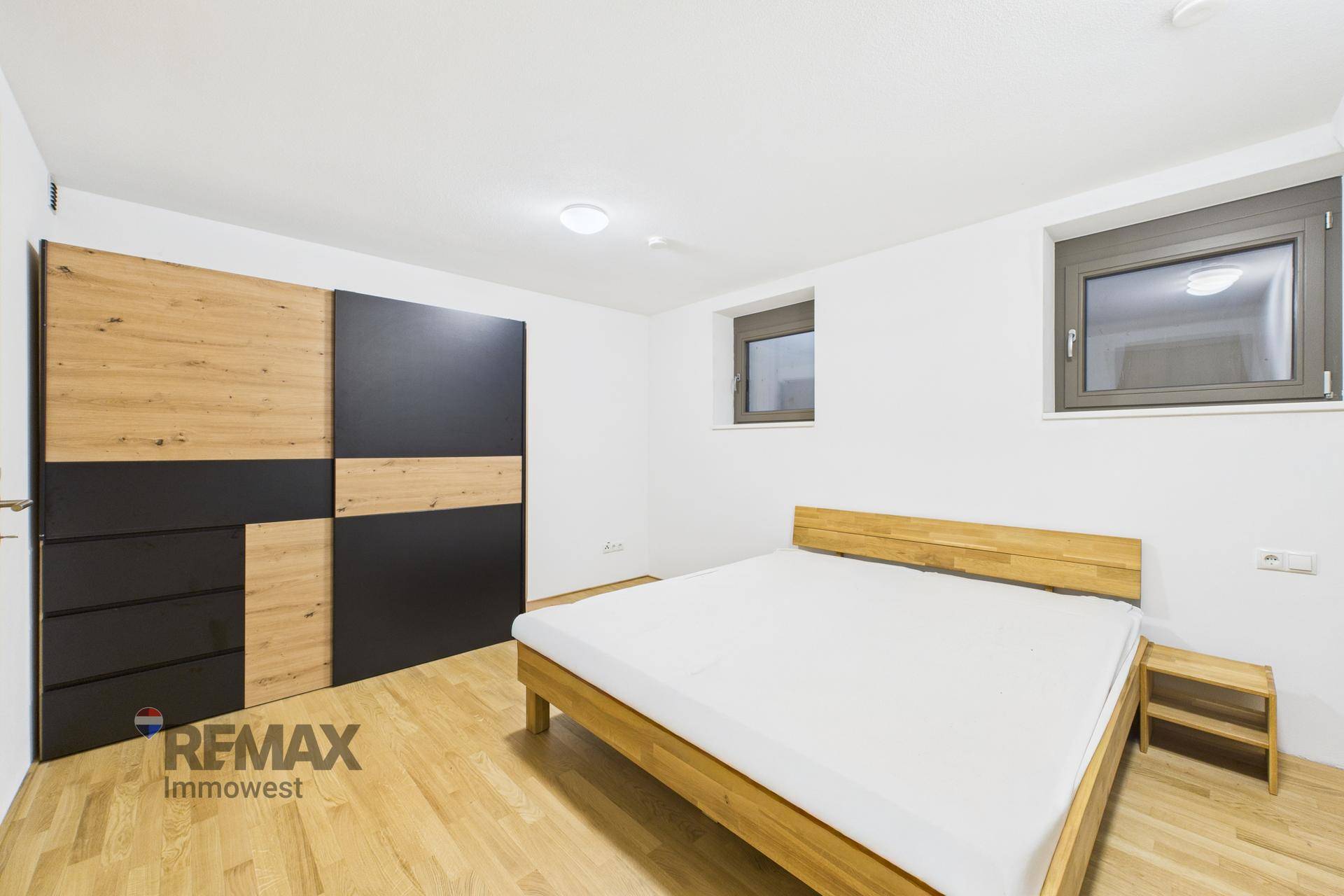 Helles Schlafzimmer mit Holzbett, modernem Kleiderschrank und zwei Fenstern für Tageslicht.