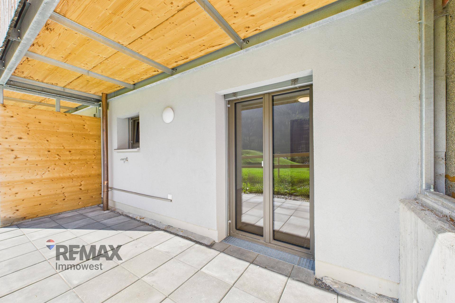 Überdachte Terrasse mit Fliesenboden und Zugang zu einem hellen Innenraum durch eine Fenstertür.