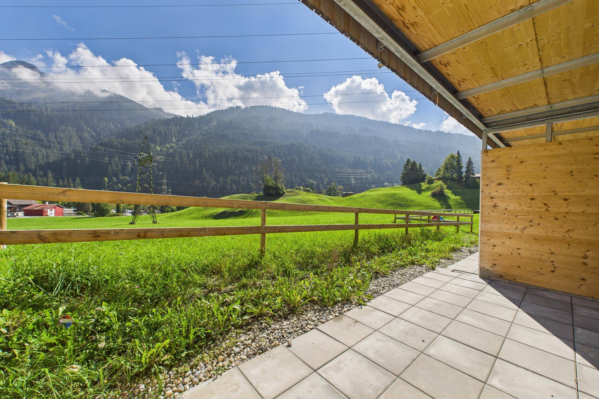 Überdachte Terrasse mit Fliesenboden und Blick auf eine grüne Wiese und die Berge.