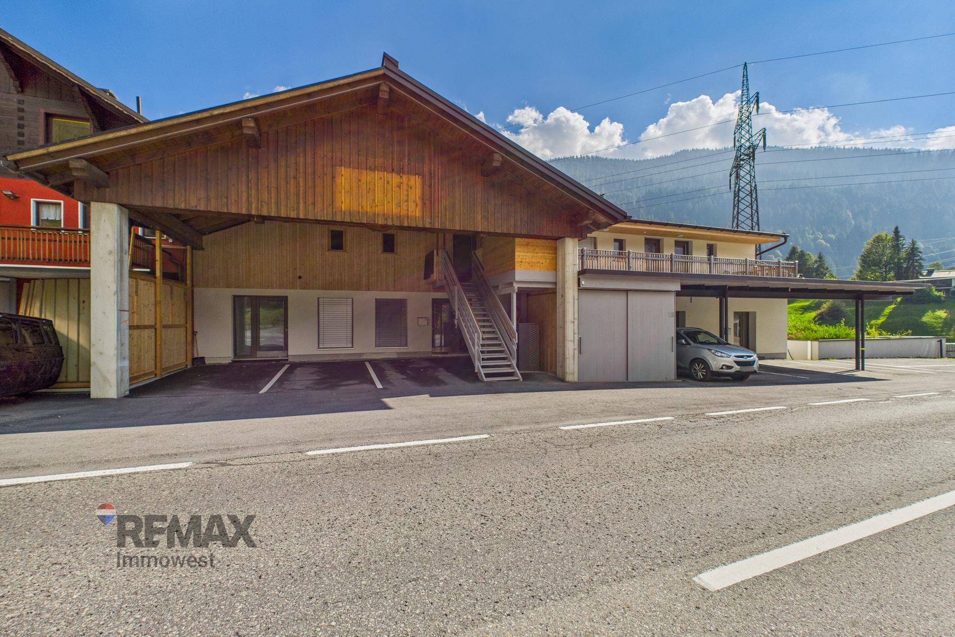 Gebäudekomplex mit Holzfassade, Carport und Parkplätzen in alpiner Umgebung.