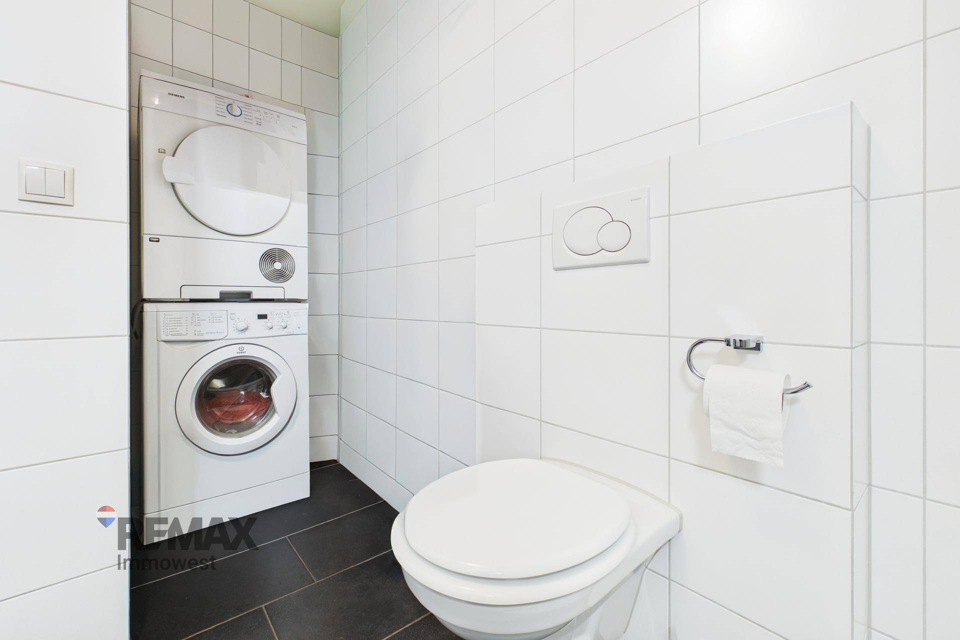 Badezimmer mit WC und integriertem Waschmaschinen- und Trocknerturm in weiß gefliestem Raum.
