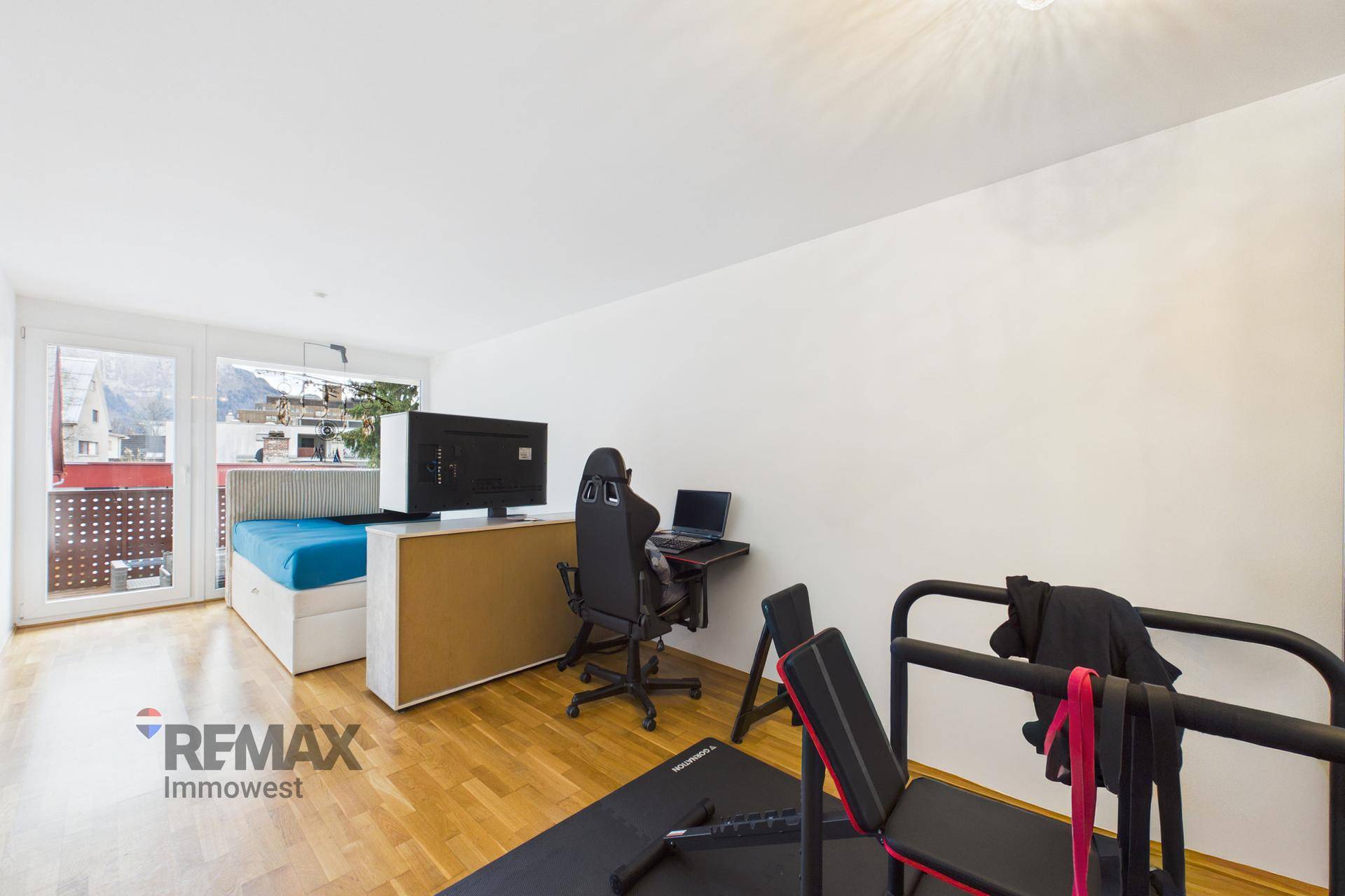 Multifunktionaler Raum mit Bett, Arbeitsbereich und Fitnessgeräten sowie Balkonzugang.